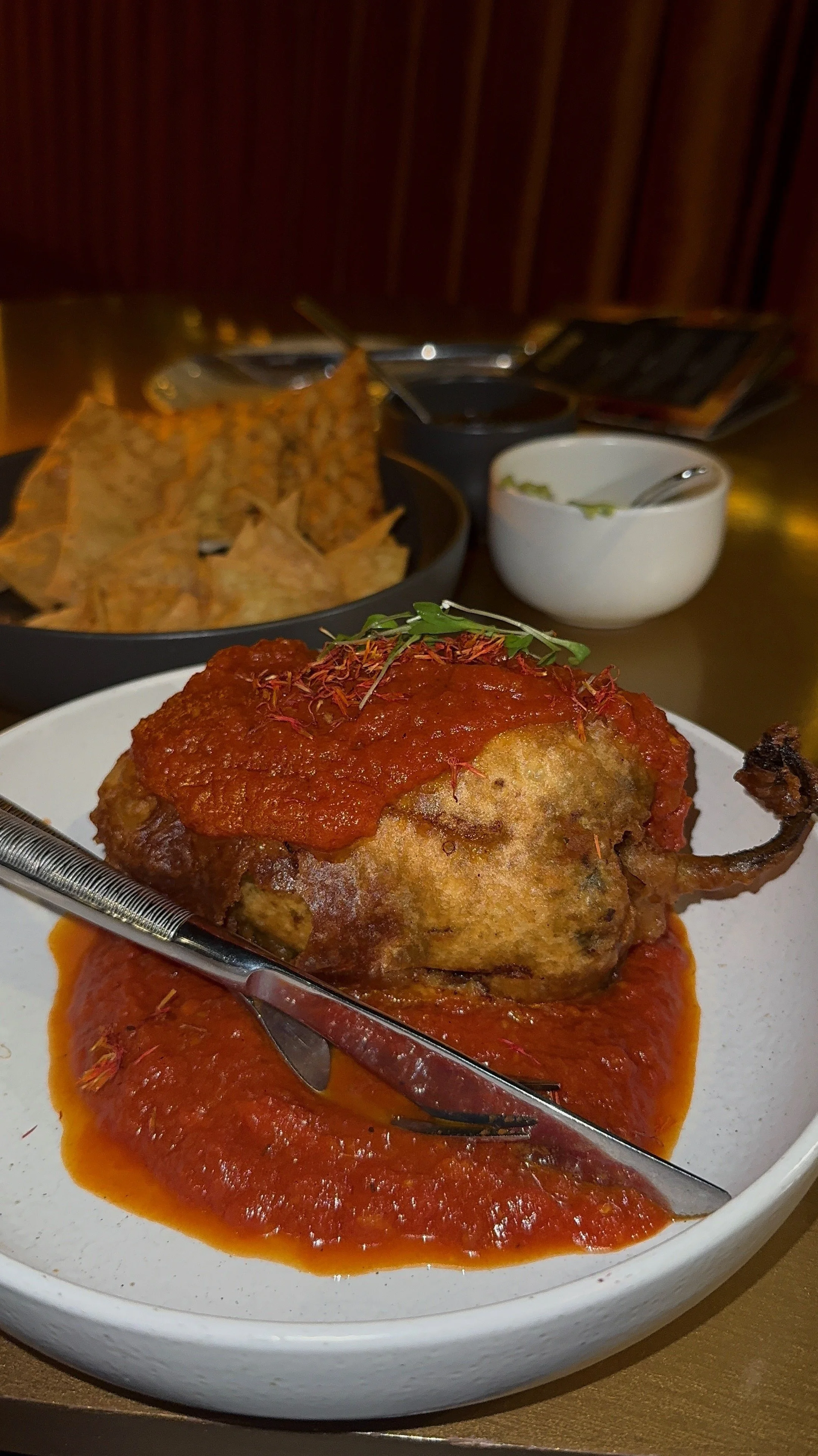 A plate with a fried chicken leg topped with tomato sauce, garnished with microgreens, served with additional sauce underneath. In the background, there are tortilla chips, a small bowl of guacamole, and a bowl of salsa, along with a cup of dark-colo