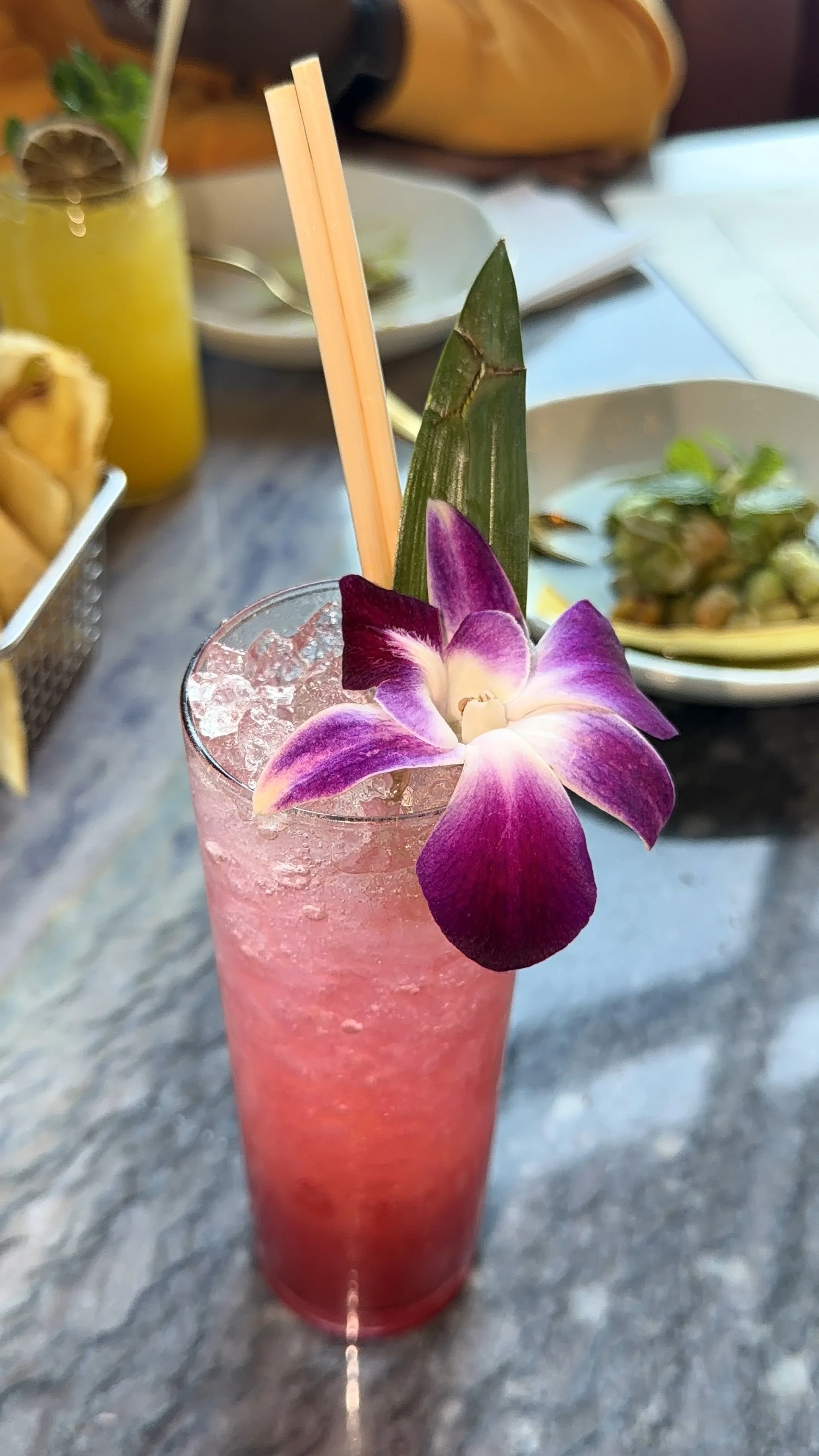 A tall glass with a pink layered cocktail garnished with an orchid flower, a pineapple leaf, and two orange straws, with ice visible inside.