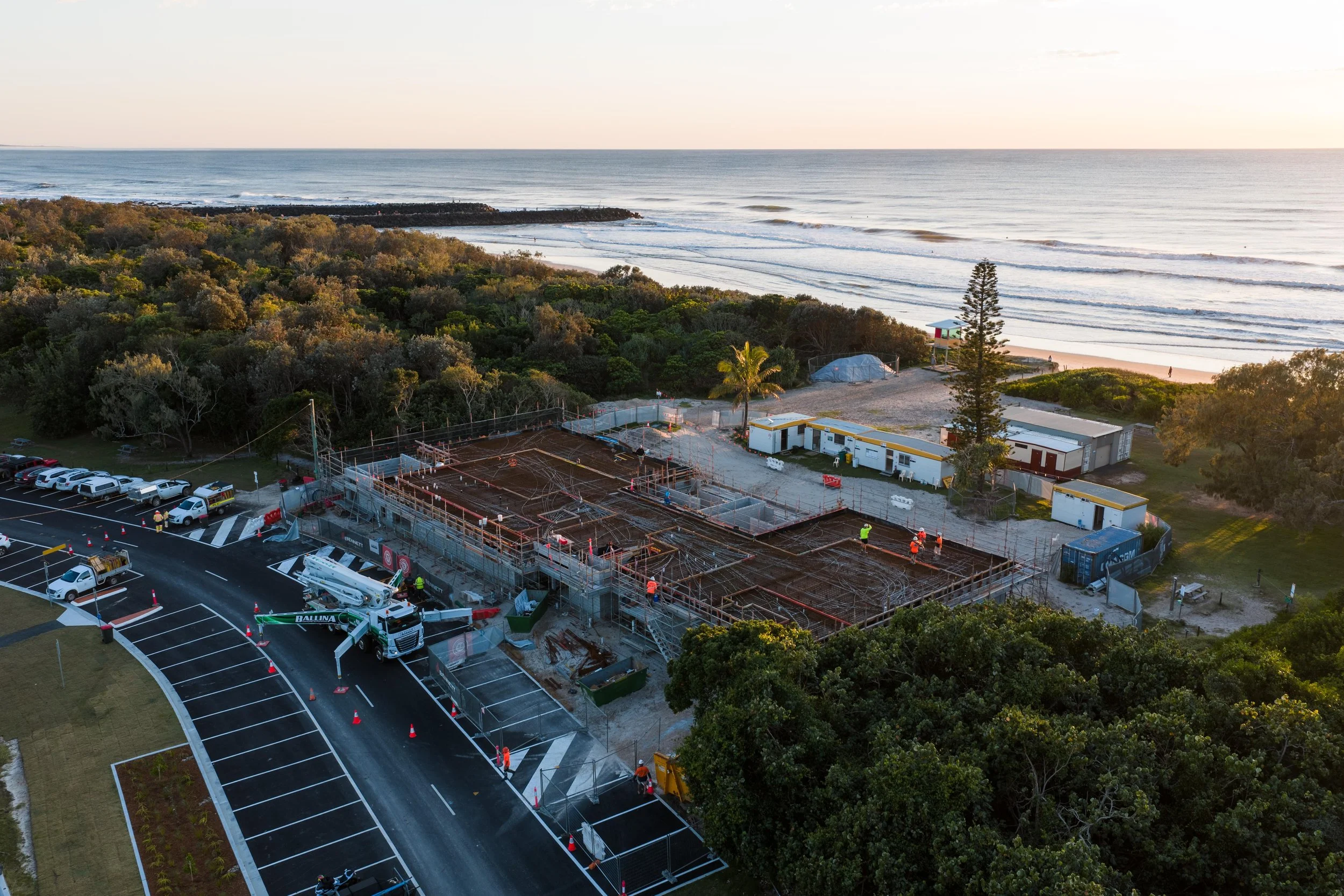 Construction site near the beach with workers, a crane, and building materials, surrounded by parking lots, trees, and ocean in the background.