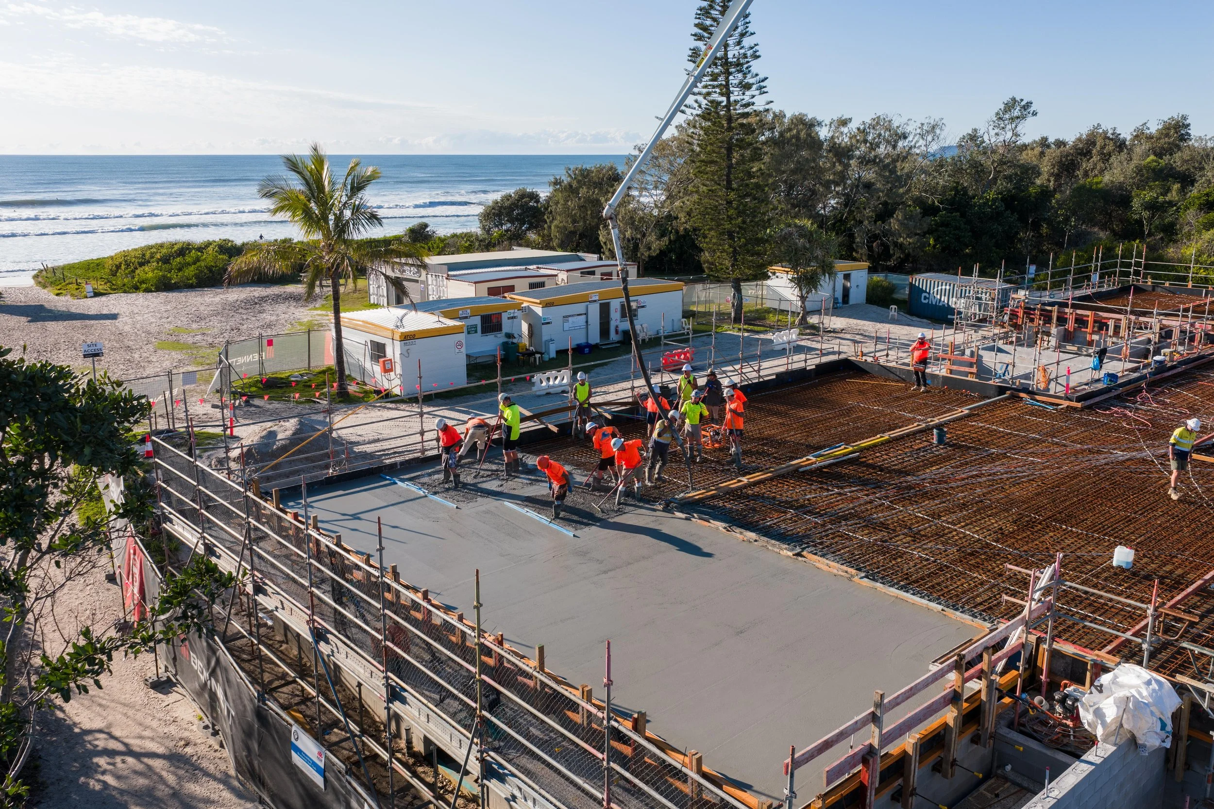 Construction workers pouring concrete for a building foundation on a beachside site with trees and ocean in the background.