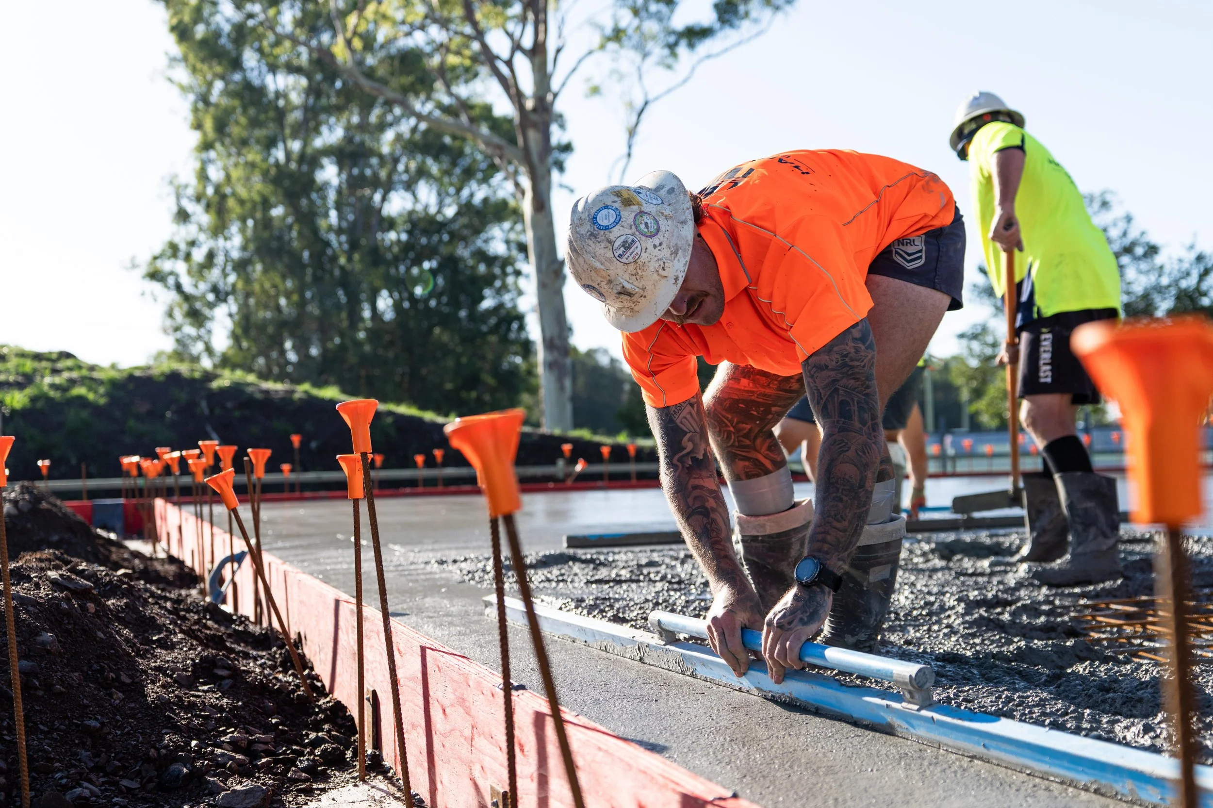 Construction workers installing concrete on a sunny outdoor site, one in an orange shirt and white helmet, the other in a yellow safety vest and helmet, with trees and a clear sky in the background.