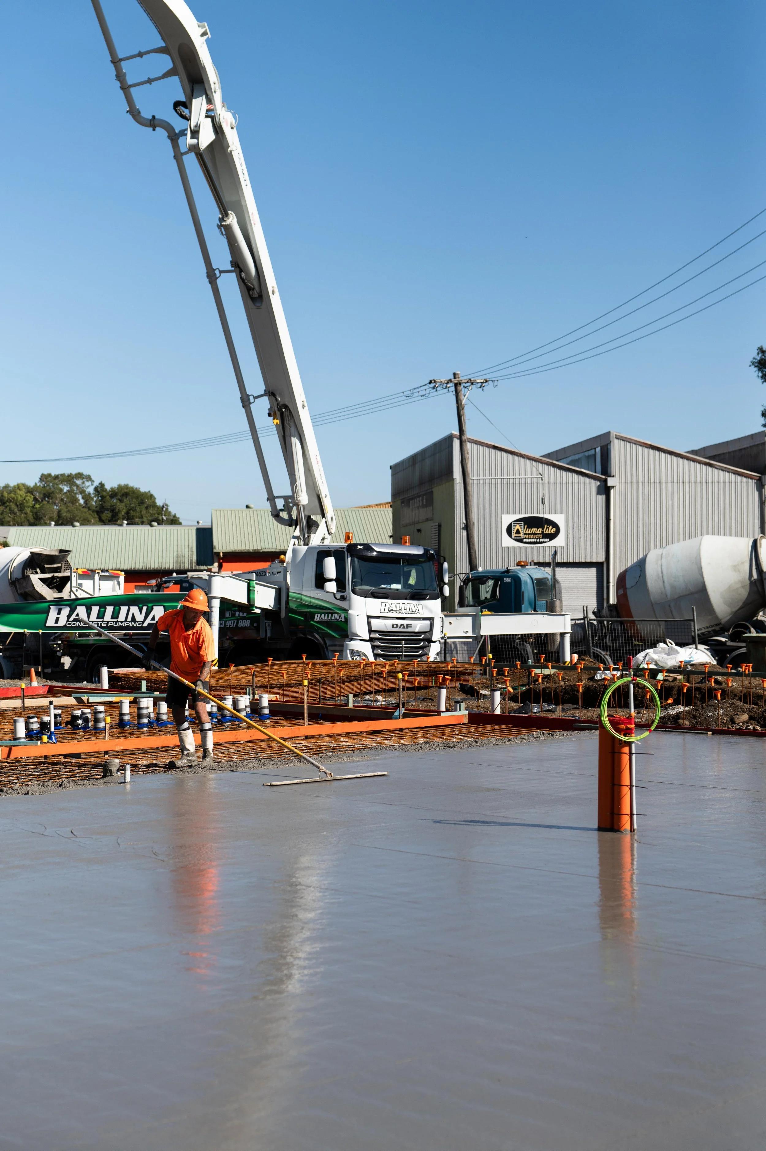 Construction worker smoothing freshly poured concrete on a building site. Construction vehicles and steel framework are visible in the background.