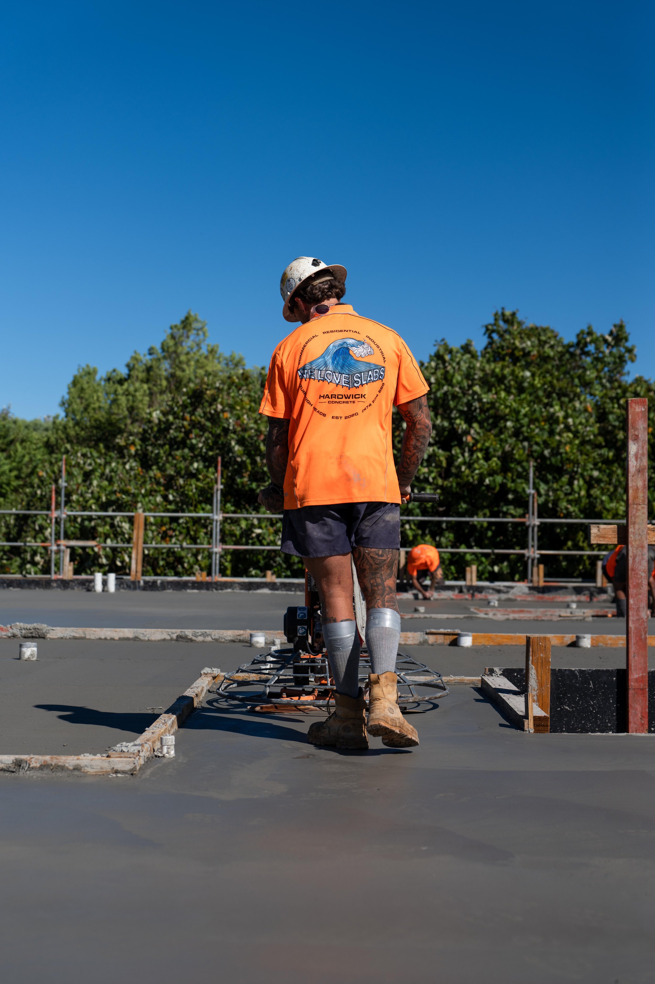A construction worker in an orange shirt and safety helmet smoothing freshly poured concrete on a building site.