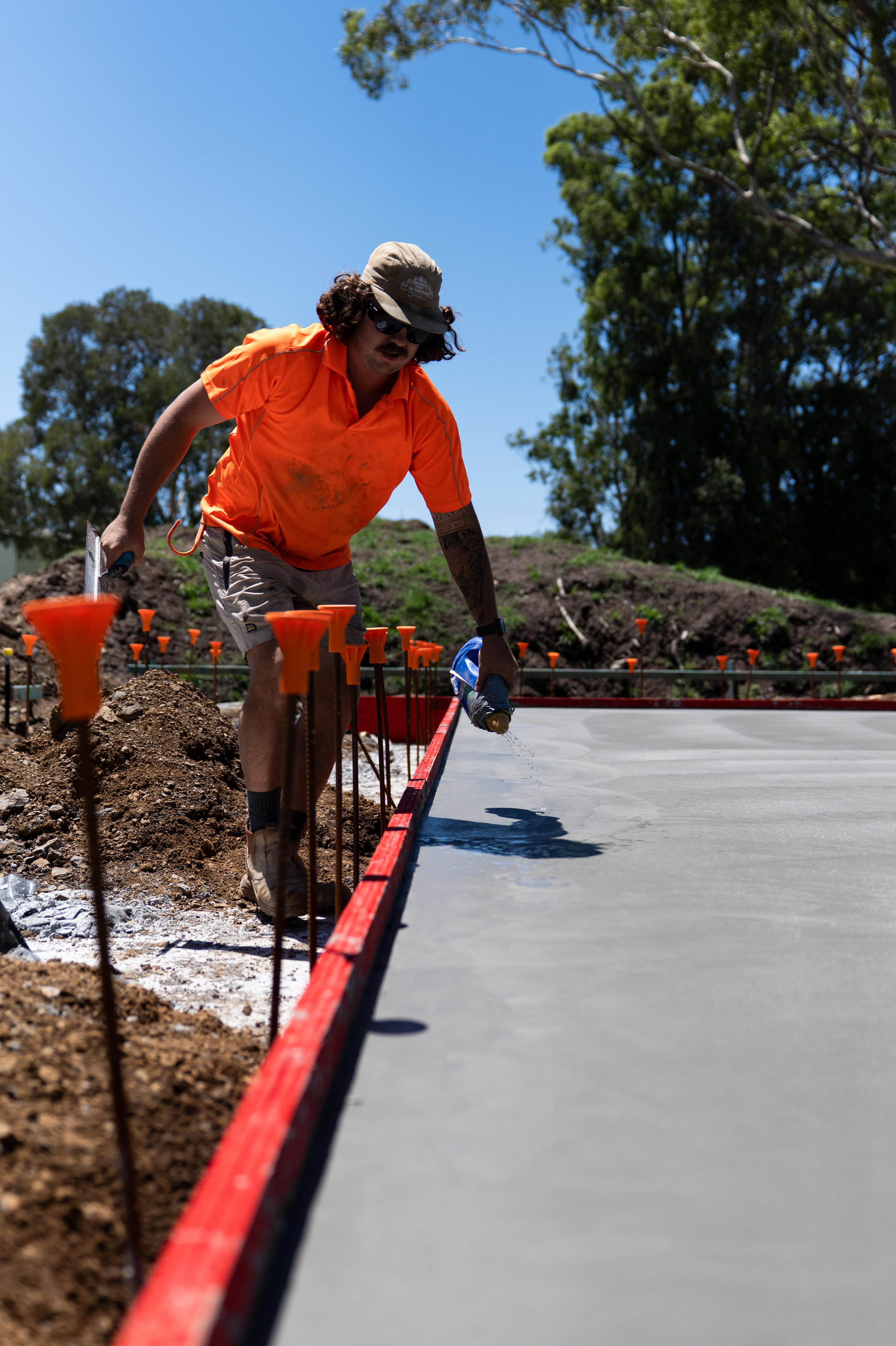 A man with long hair, sunglasses, and a hat spray-paints a concrete surface at an outdoor construction site on a sunny day.