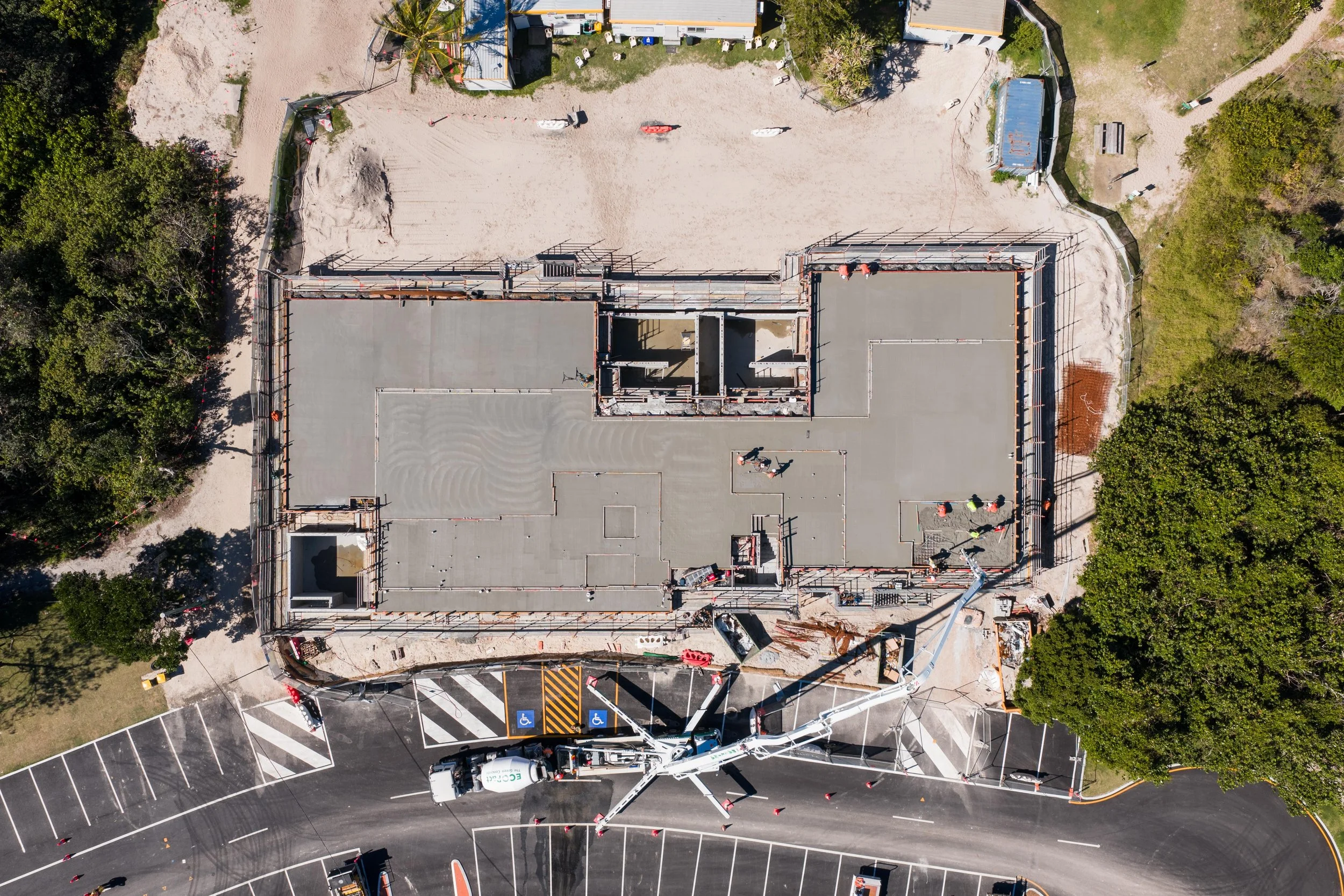 An aerial view of a construction site with a building in progress, surrounded by trees, parking lot, and construction equipment.