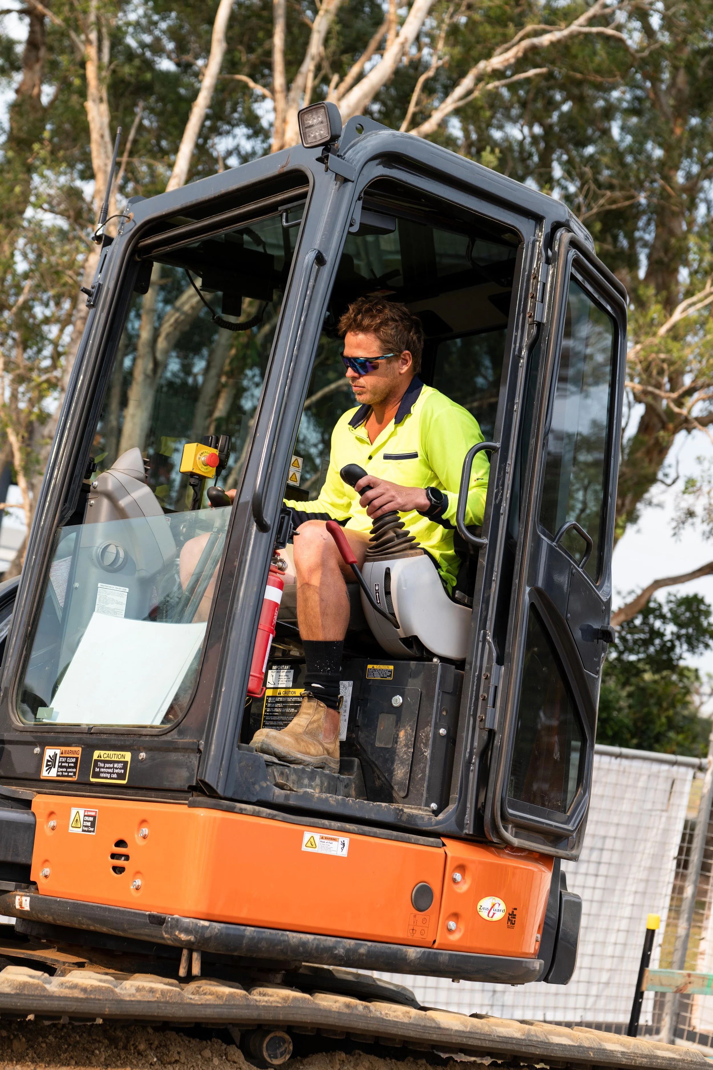 A man wearing sunglasses, a fluorescent yellow safety shirt, and tan boots operating an orange and black excavator with a glass enclosed cab, on a construction site with trees in the background.