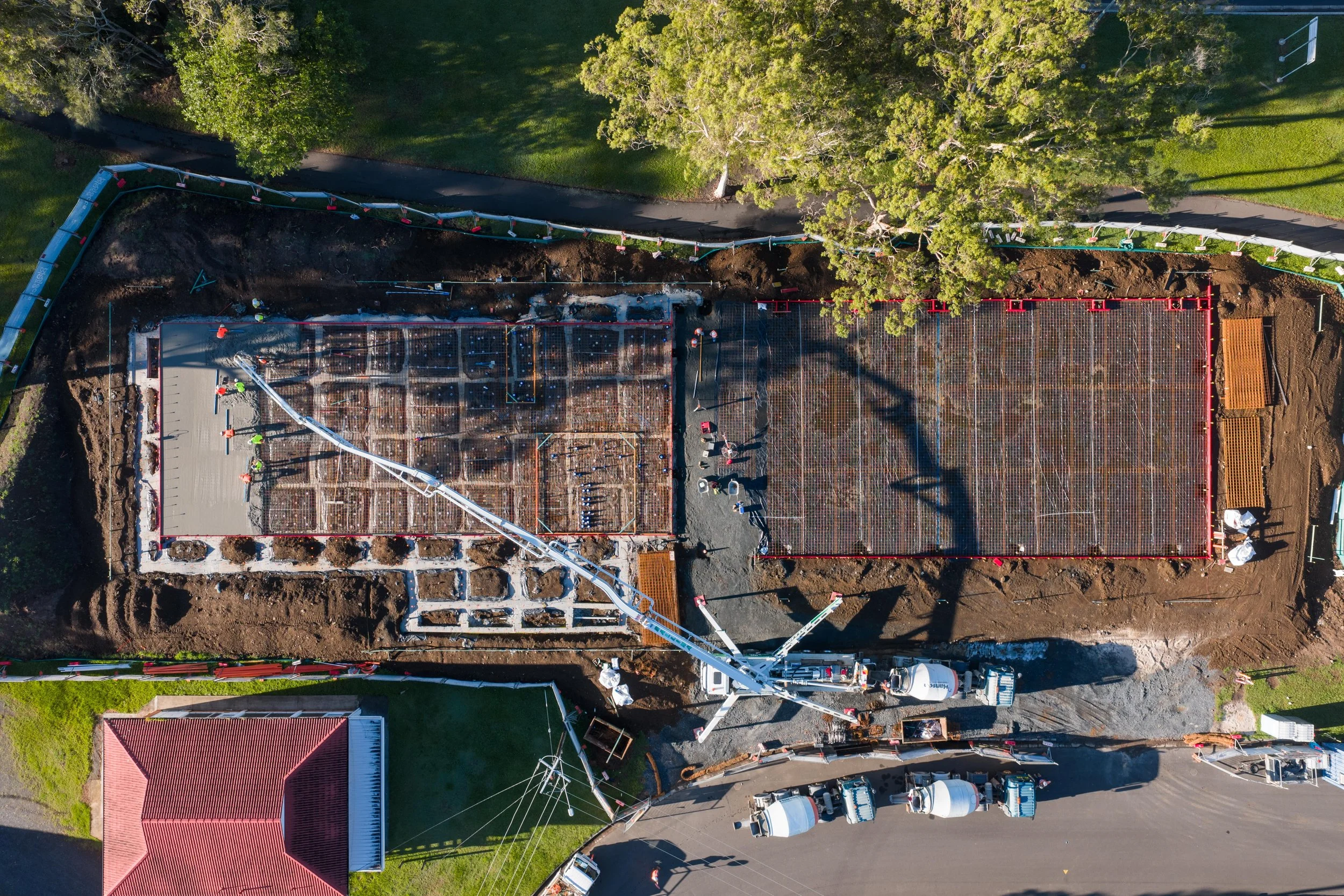 An aerial view of a construction site showing two large concrete foundations with rebar and framing. Workers and construction equipment are present, with a crane and cement trucks nearby. The site is surrounded by trees, a road, and residential house