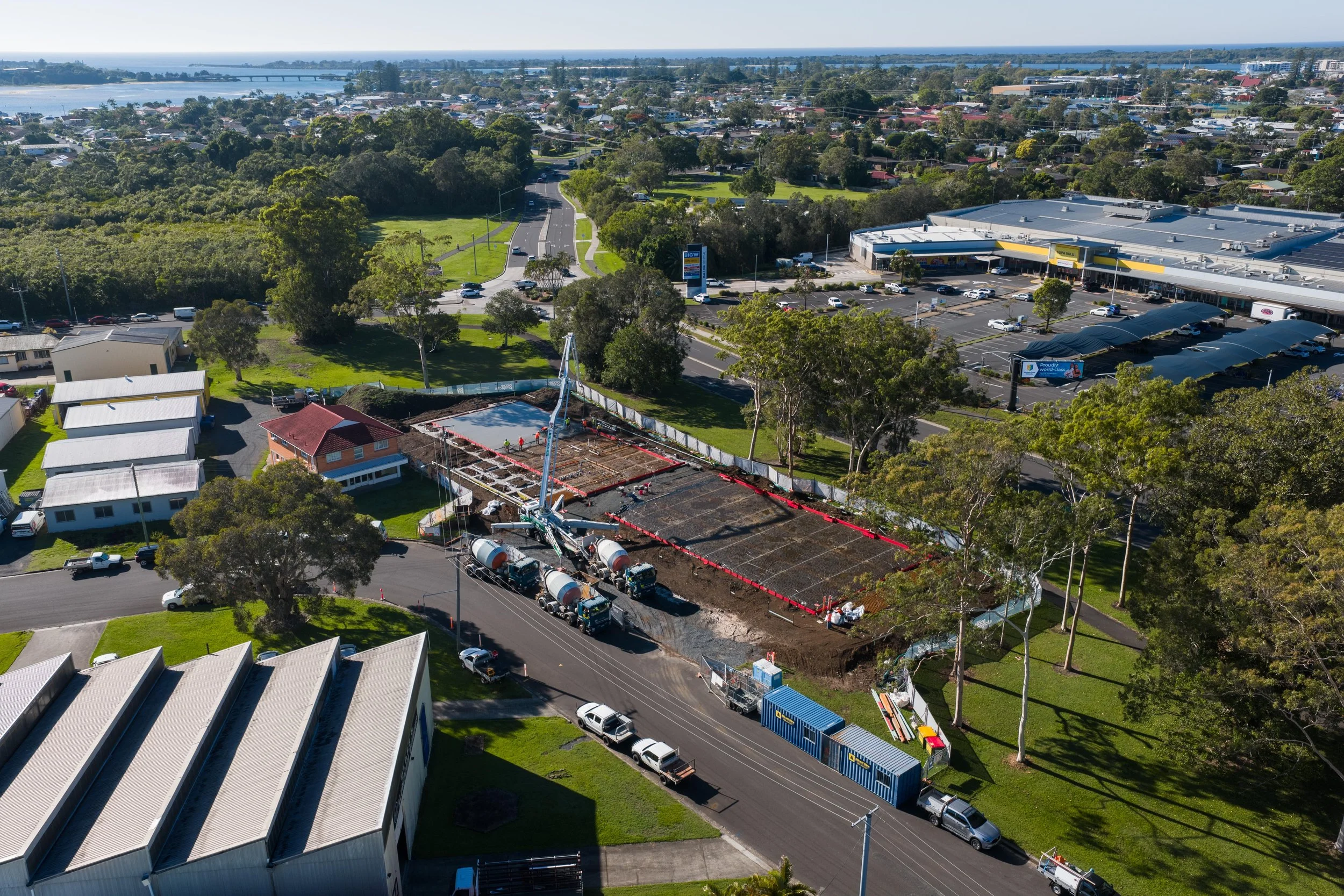 Construction site with cement mixers and foundation work, surrounded by trees, residential houses, and commercial area in the background.