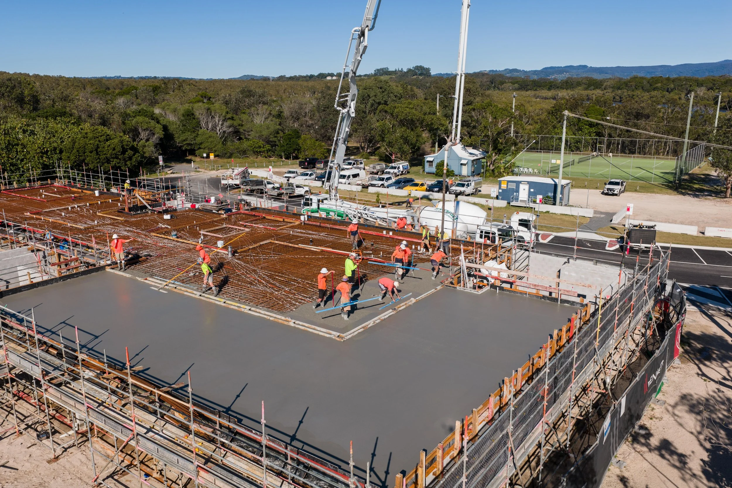 Construction workers in orange and yellow shirts work on pouring and leveling concrete on a building foundation, with construction equipment and vehicles visible around the site.