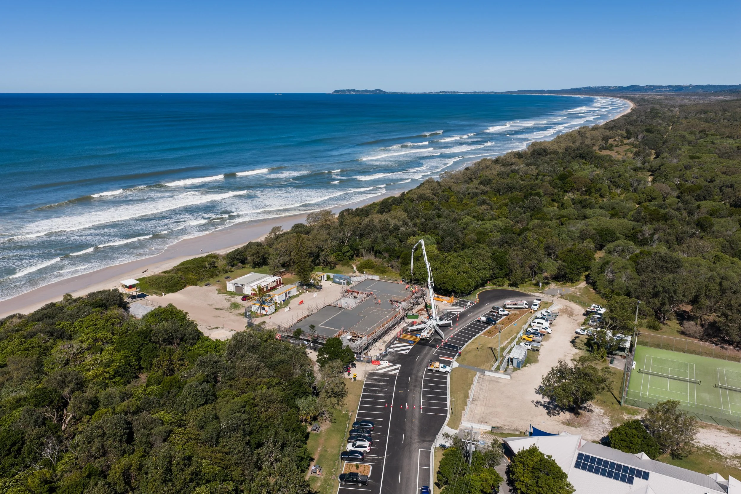 An aerial view of a sunny beach with waves, a parking lot with cars, a fenced sports court, and a large construction crane set up at a park near the coastline, surrounded by green trees.