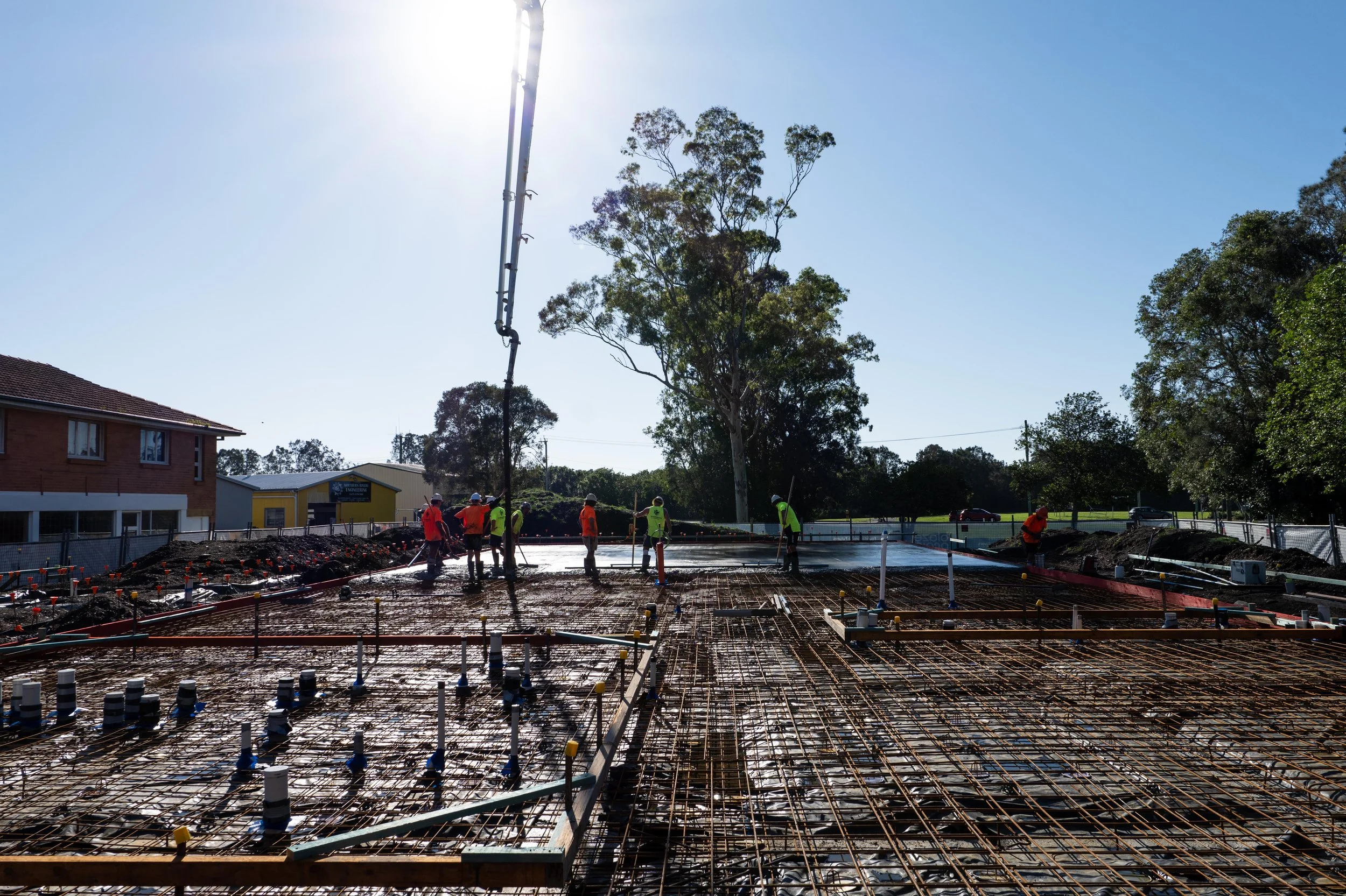 Construction workers pouring concrete on a building foundation at a construction site with steel rebar reinforcement, trees, and buildings in the background.