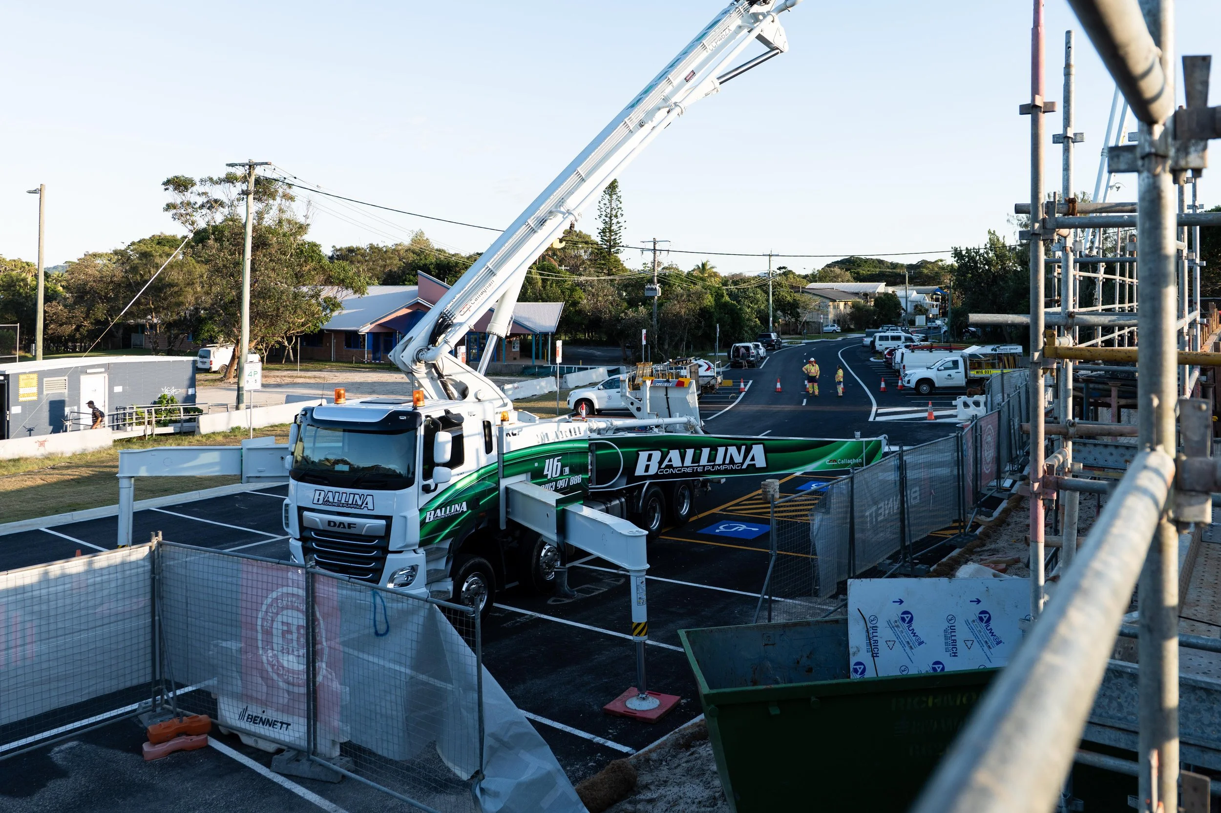 Construction site with a large green and white concrete pumping truck labeled 'Ballina' in a parking lot, with workers in safety gear, fencing, and scaffolding.