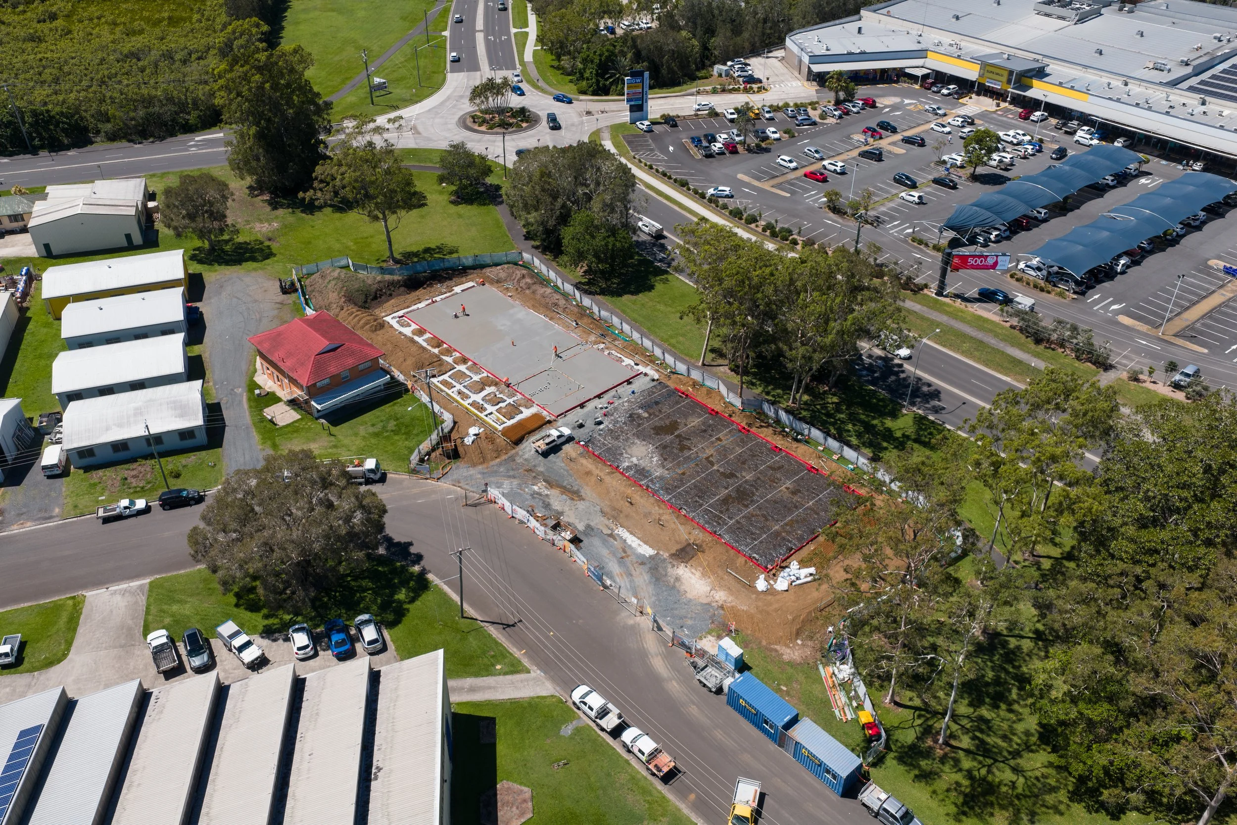 Aerial view of a construction site with two partially poured concrete slabs, near a busy shopping center parking lot. The site is fenced, with construction equipment and workers present. Surrounding area includes parking lots, green spaces, and residential buildings.