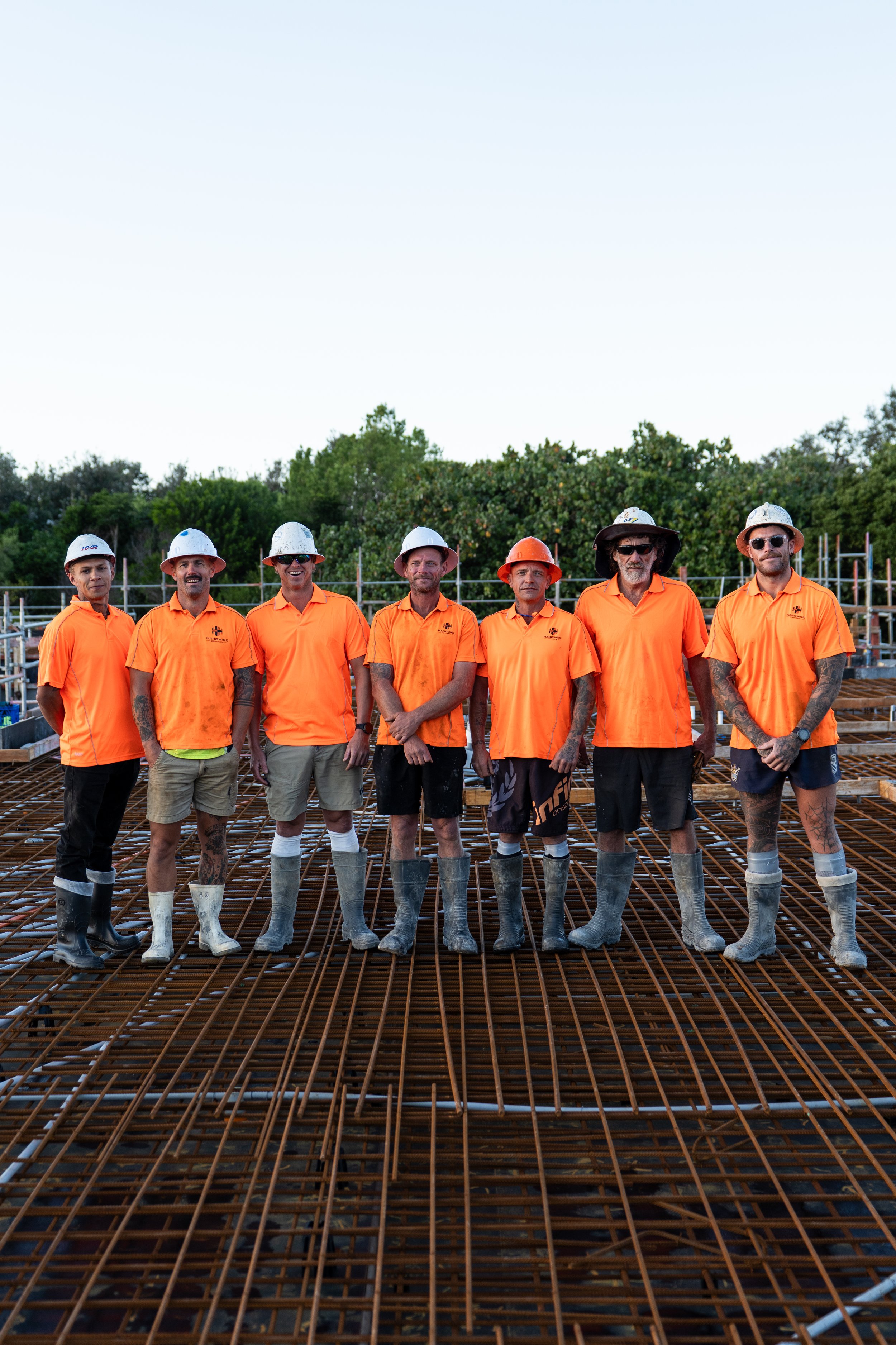 Seven construction workers stand in a row on a steel rebar framework at a construction site during daytime, wearing orange shirts, safety helmets, and work boots with a backdrop of trees and a clear sky.