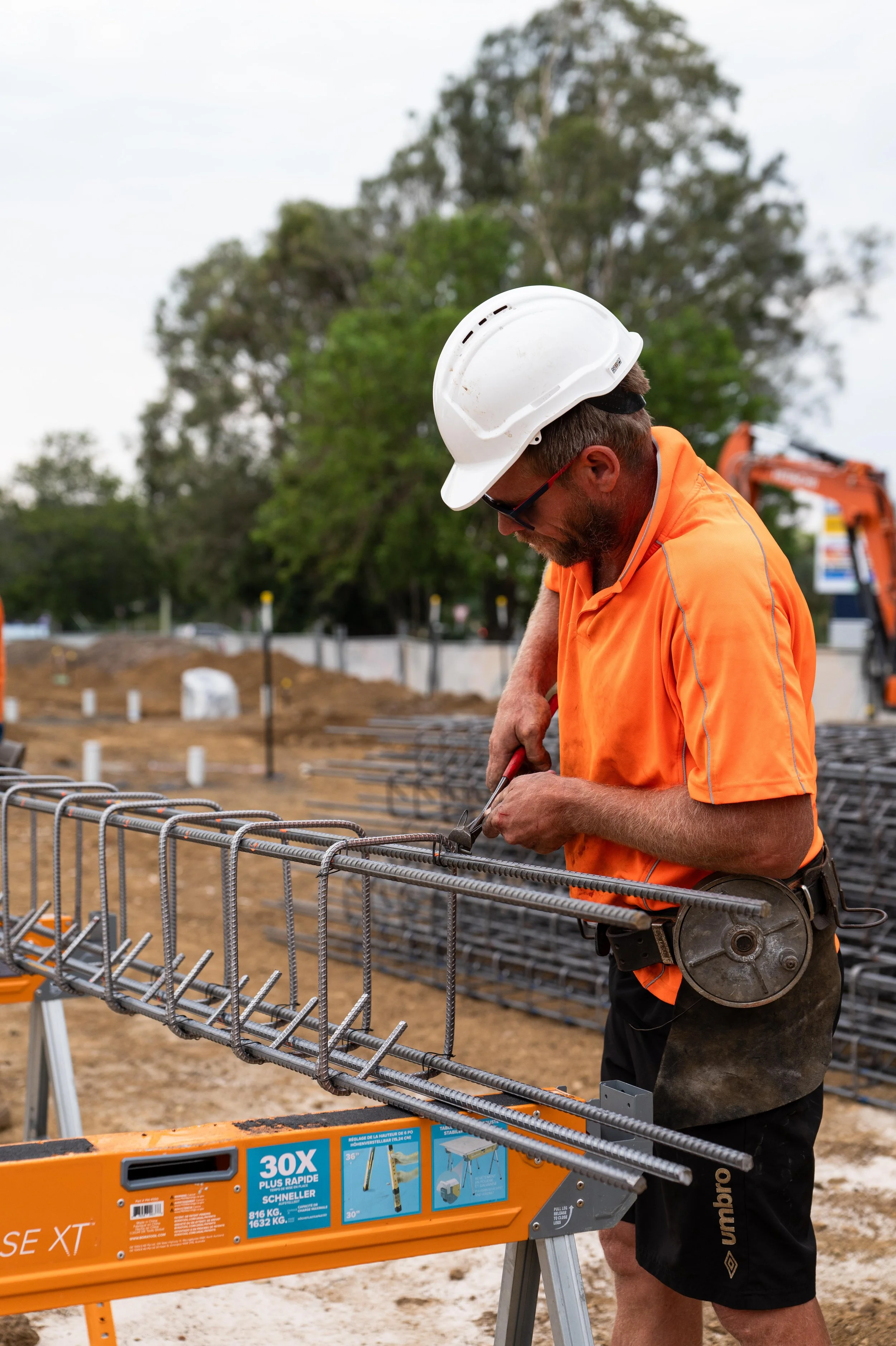 A construction worker in an orange shirt, white helmet, and sunglasses working on steel rebar at a construction site, with trees and construction equipment in the background.