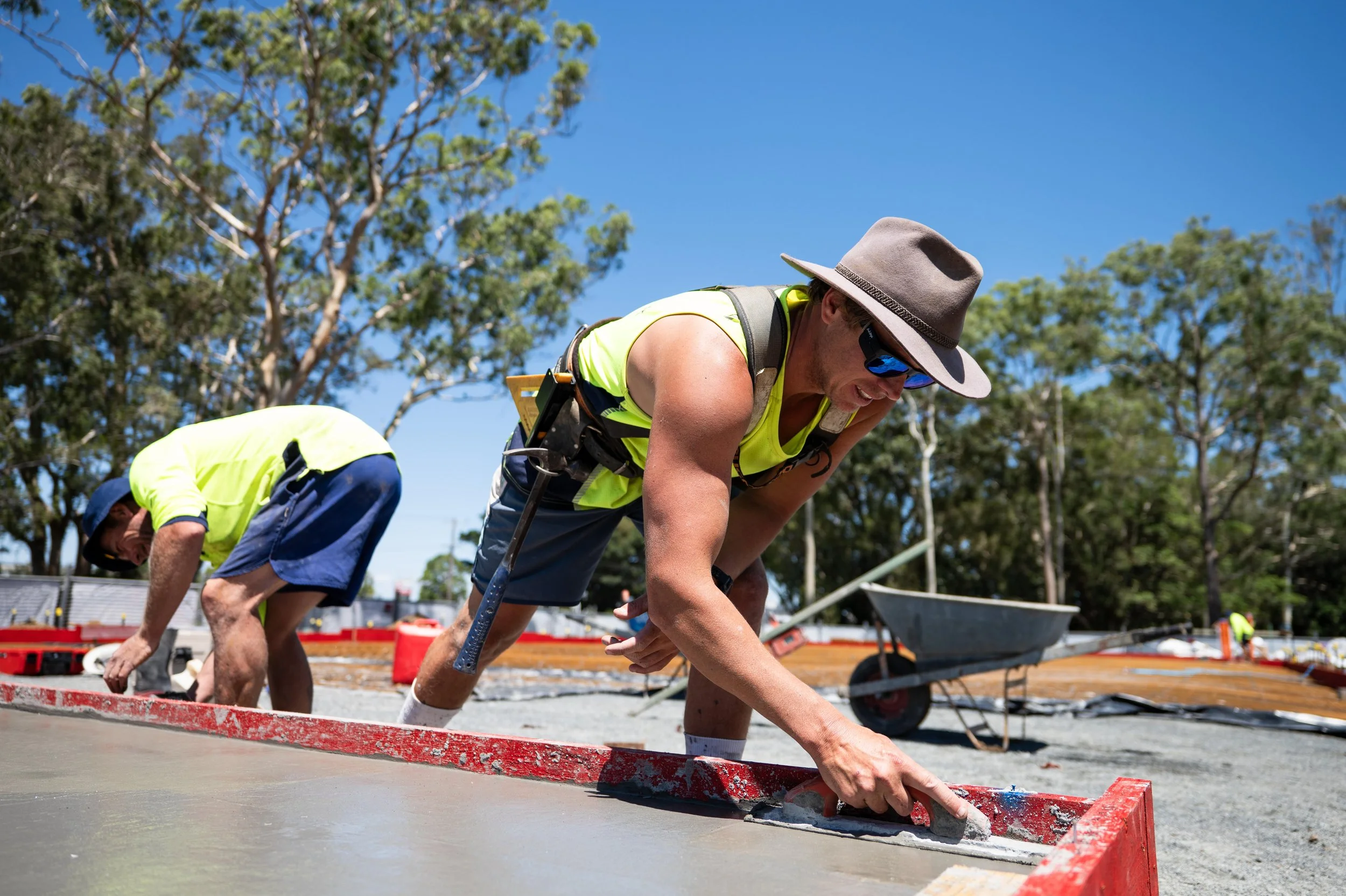 Two construction workers in fluorescent yellow safety shirts and hats pouring and smoothing concrete on a construction site with trees and blue sky in the background.