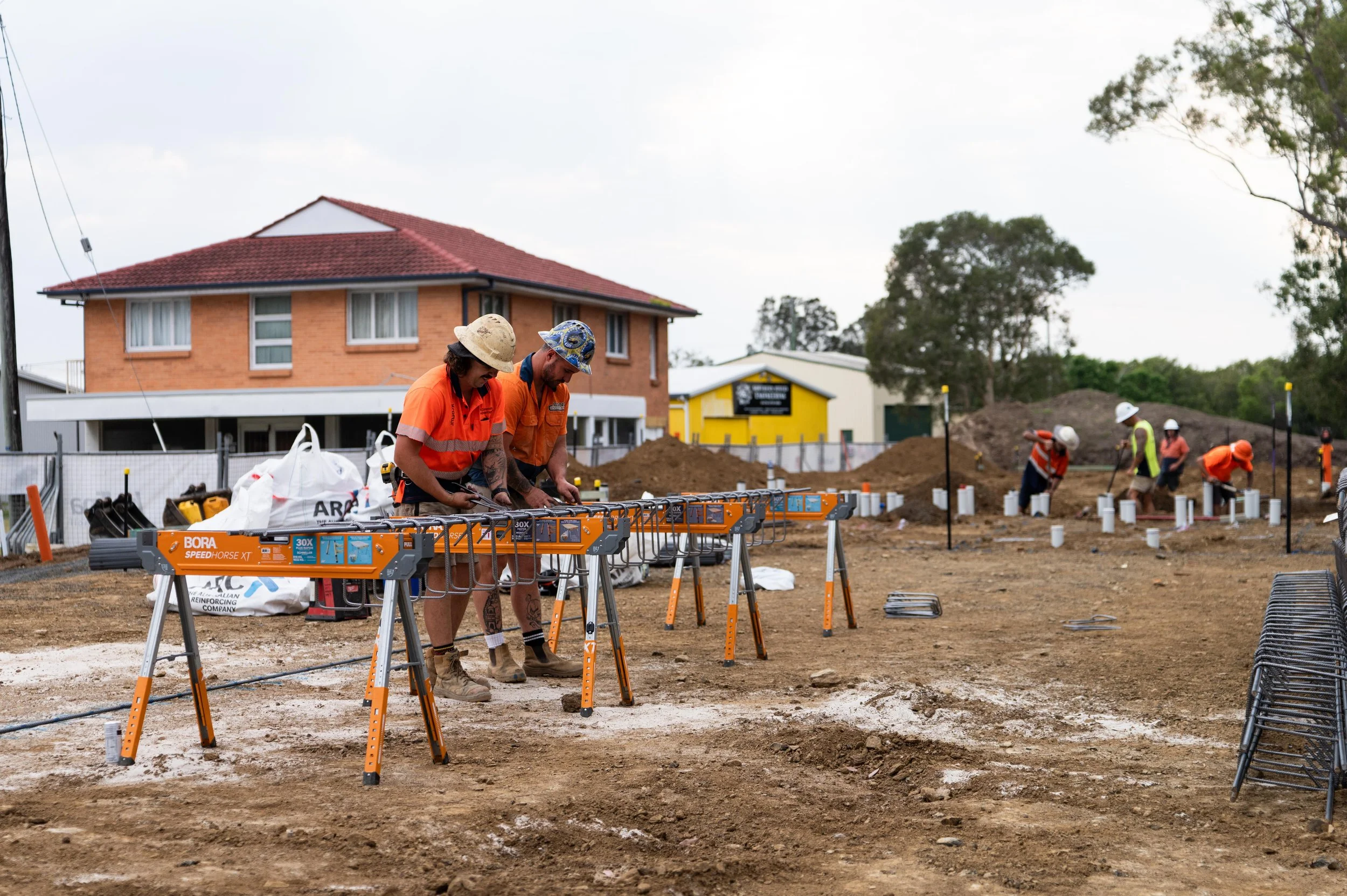 Construction workers working on a construction site with building materials and tools, some workers are working with machinery, and others are digging or preparing ground for construction.