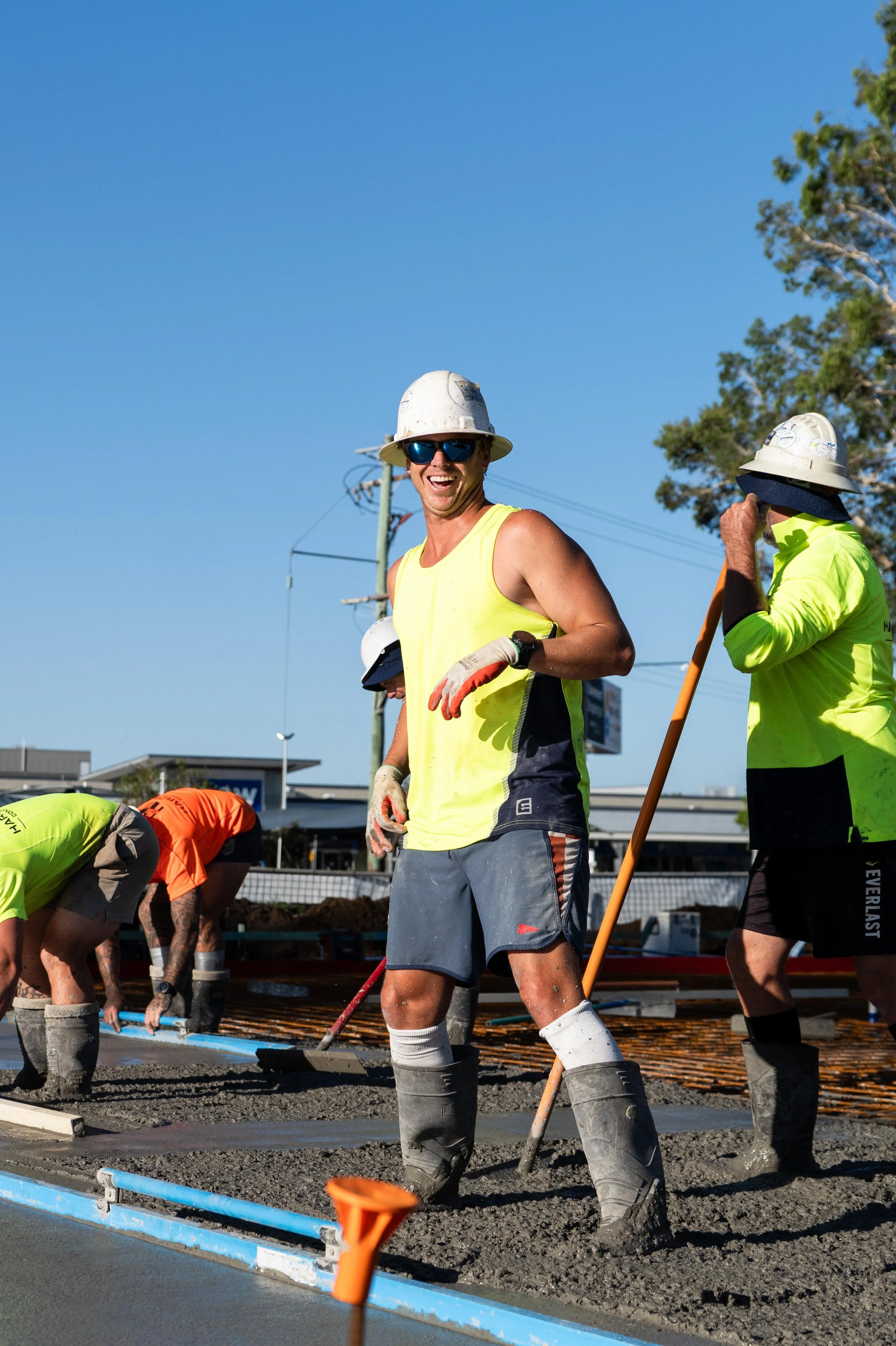 Construction workers, wearing safety helmets and reflective vests, are pouring concrete on a construction site under a clear blue sky.