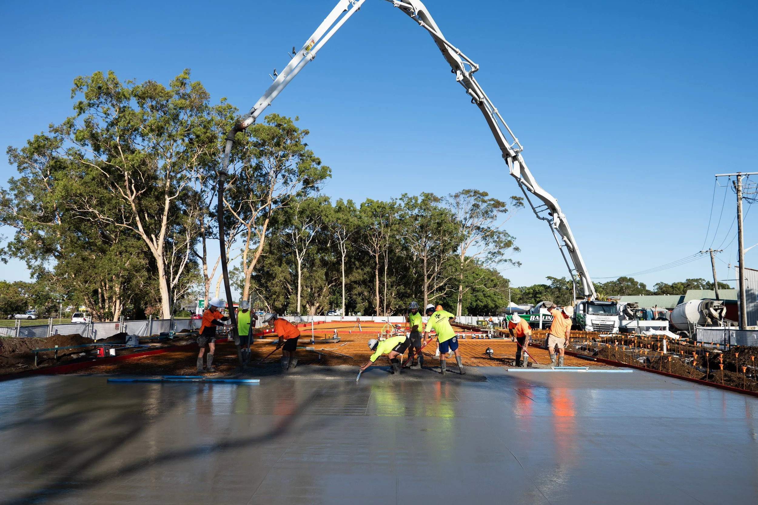 Construction workers in brightly colored shirts and hard hats pouring and spreading wet concrete on a construction site with trees and utility poles in the background.