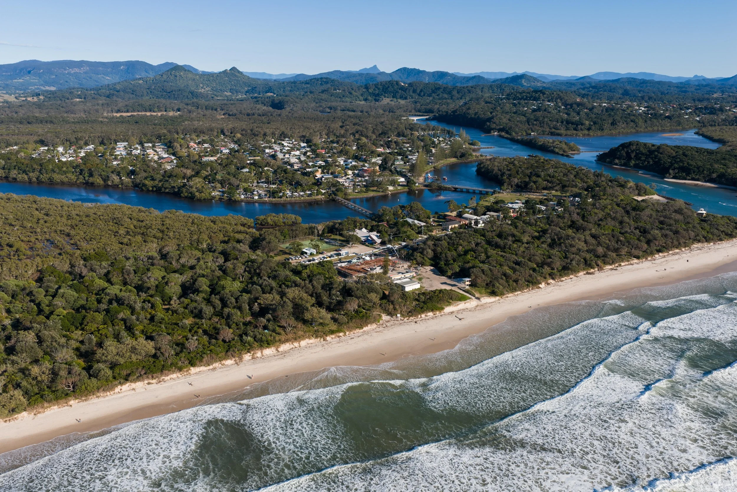 Aerial view of a beach with dunes and waves, a forested area, a river with boats, and a town with houses and infrastructure against a mountain backdrop.