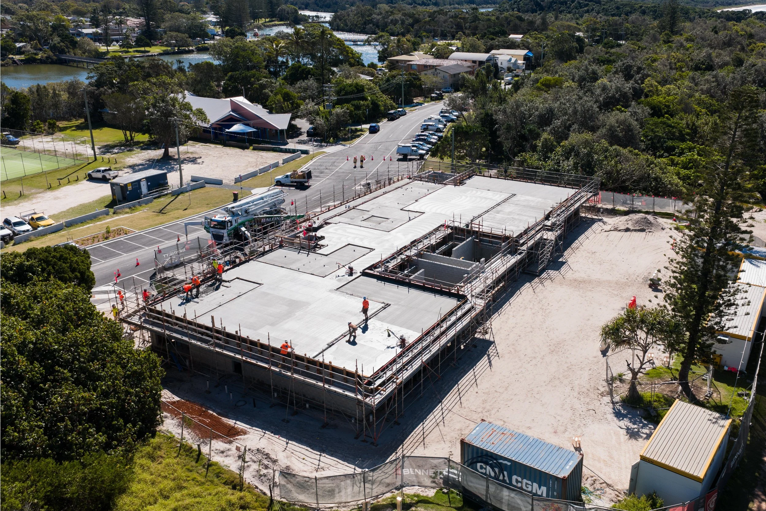 Aerial view of a construction site showing workers on a concrete foundation, surrounded by scaffolding, with a parking lot, trees, and water bodies in the background.