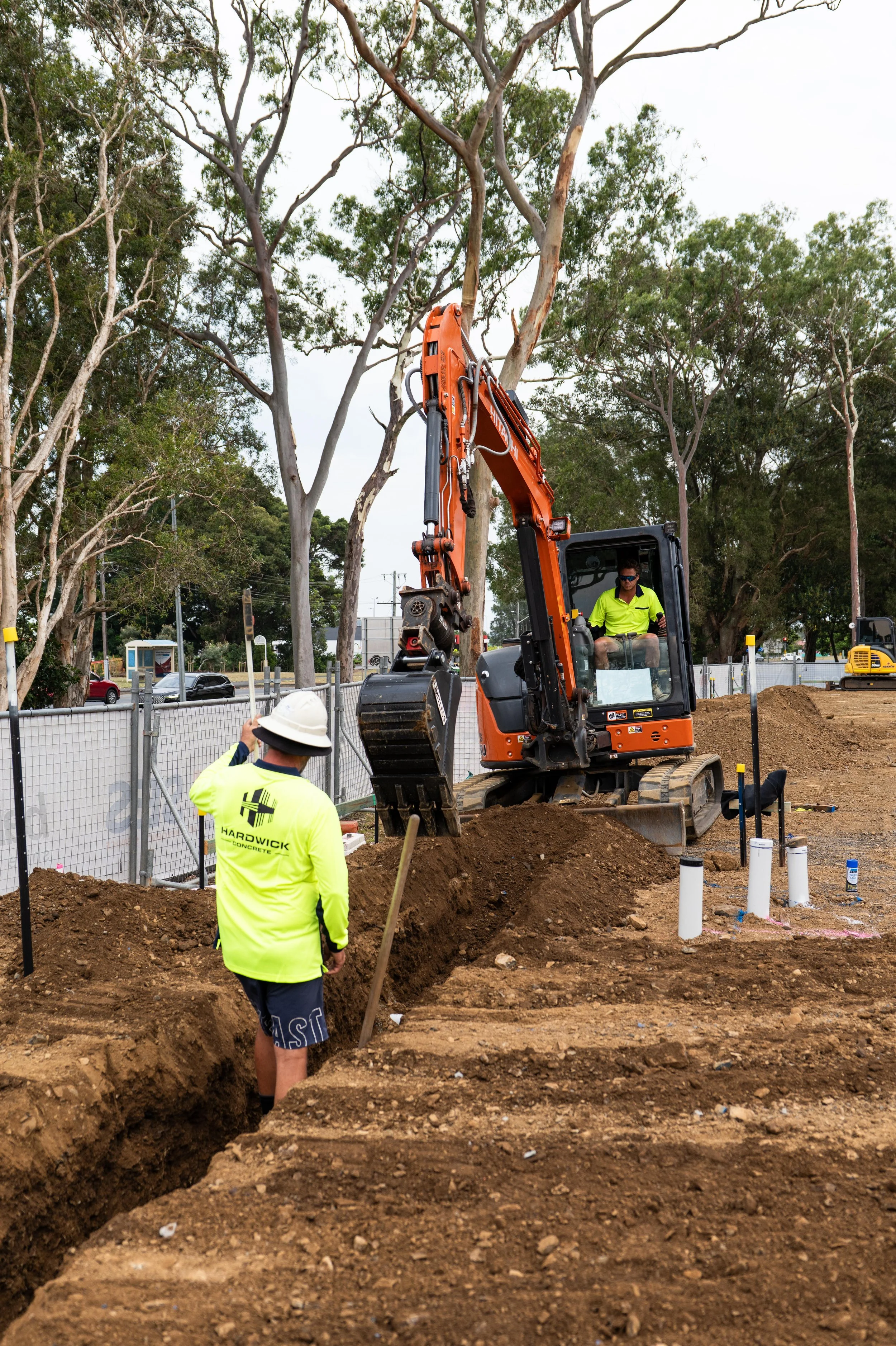 Construction workers and an orange excavator at a construction site with dirt trenches, pipes, and trees in the background.