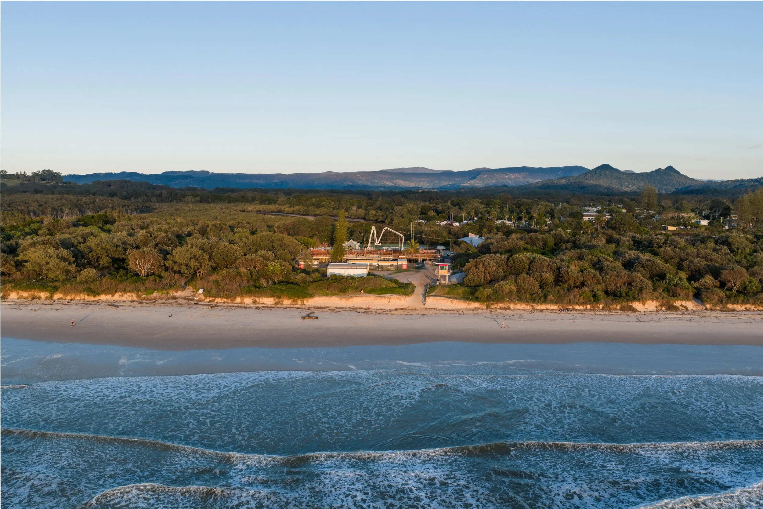 Aerial view of a beach with ocean waves in the foreground, a sandy shore, green shrubbery, and a amusement park with rides in the background, set against distant mountains under a clear sky.