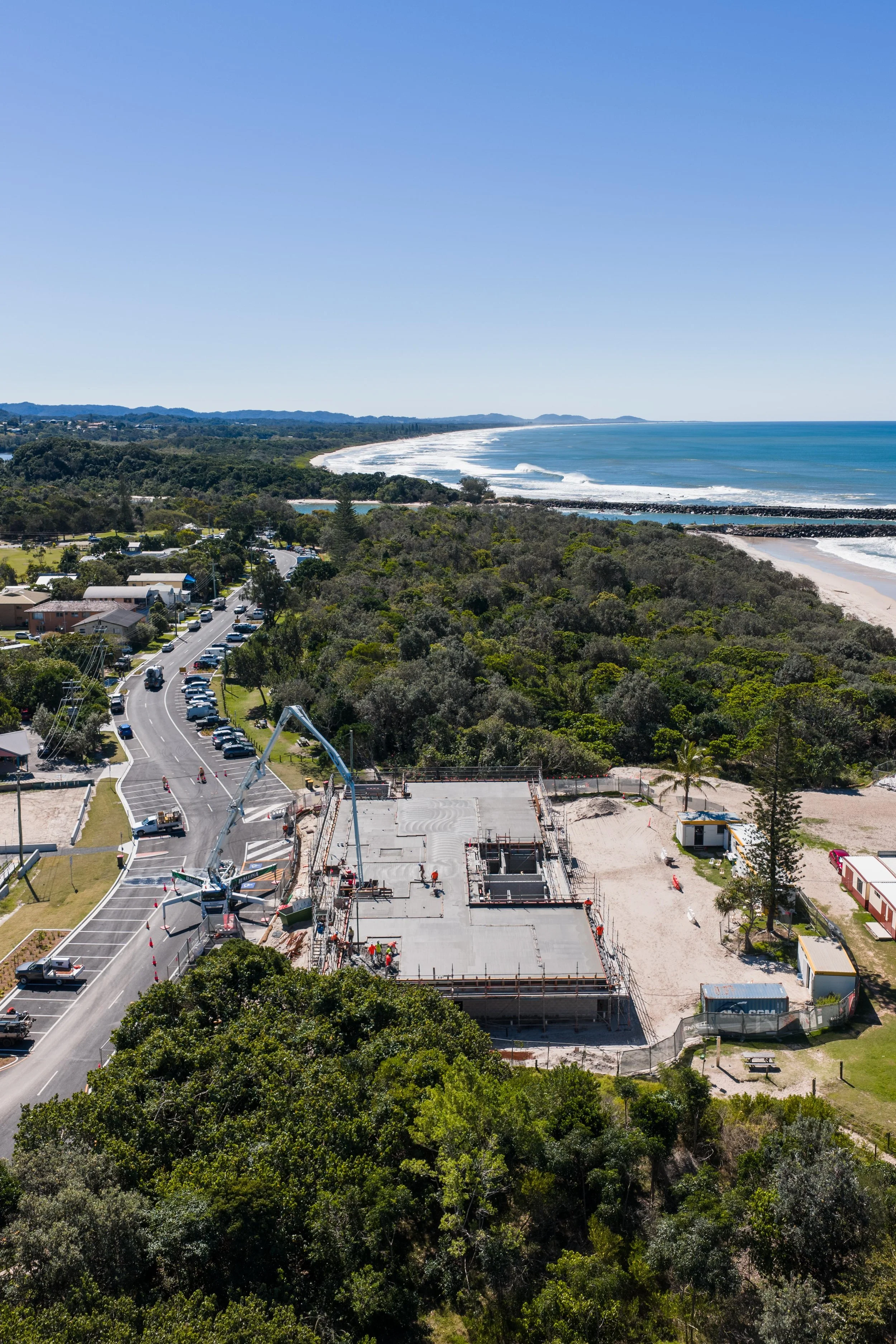 An aerial view of a construction site near a coastline with trees, a road, and parked cars, and the ocean in the background.