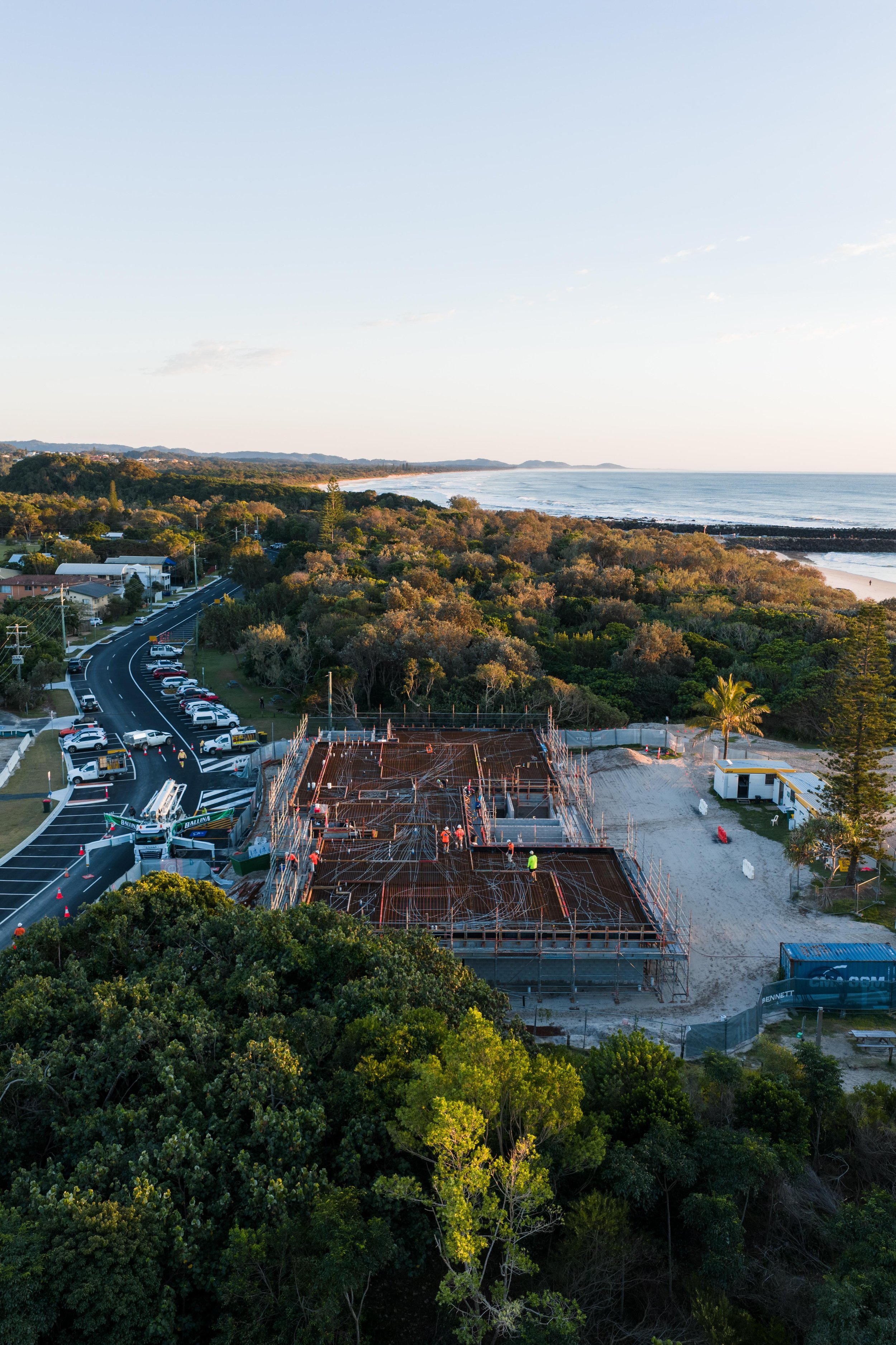 Construction site on a beach with workers, scaffolding, and equipment, near a wooded area and ocean, under clear skies.