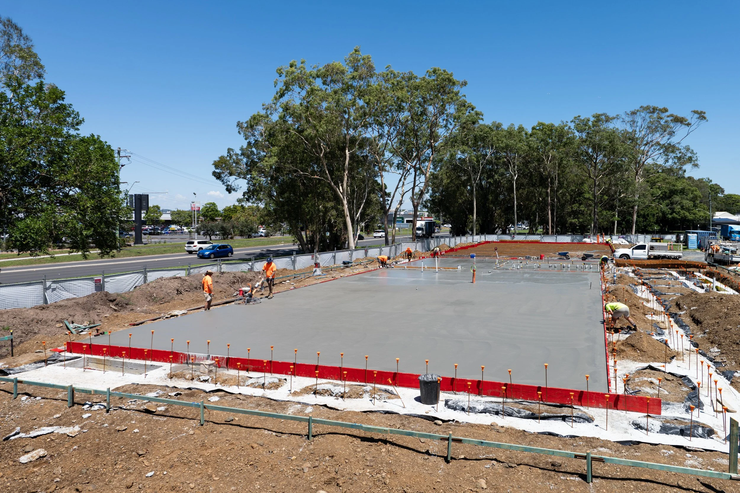 Construction workers pouring concrete on a building foundation with trees and traffic in the background.