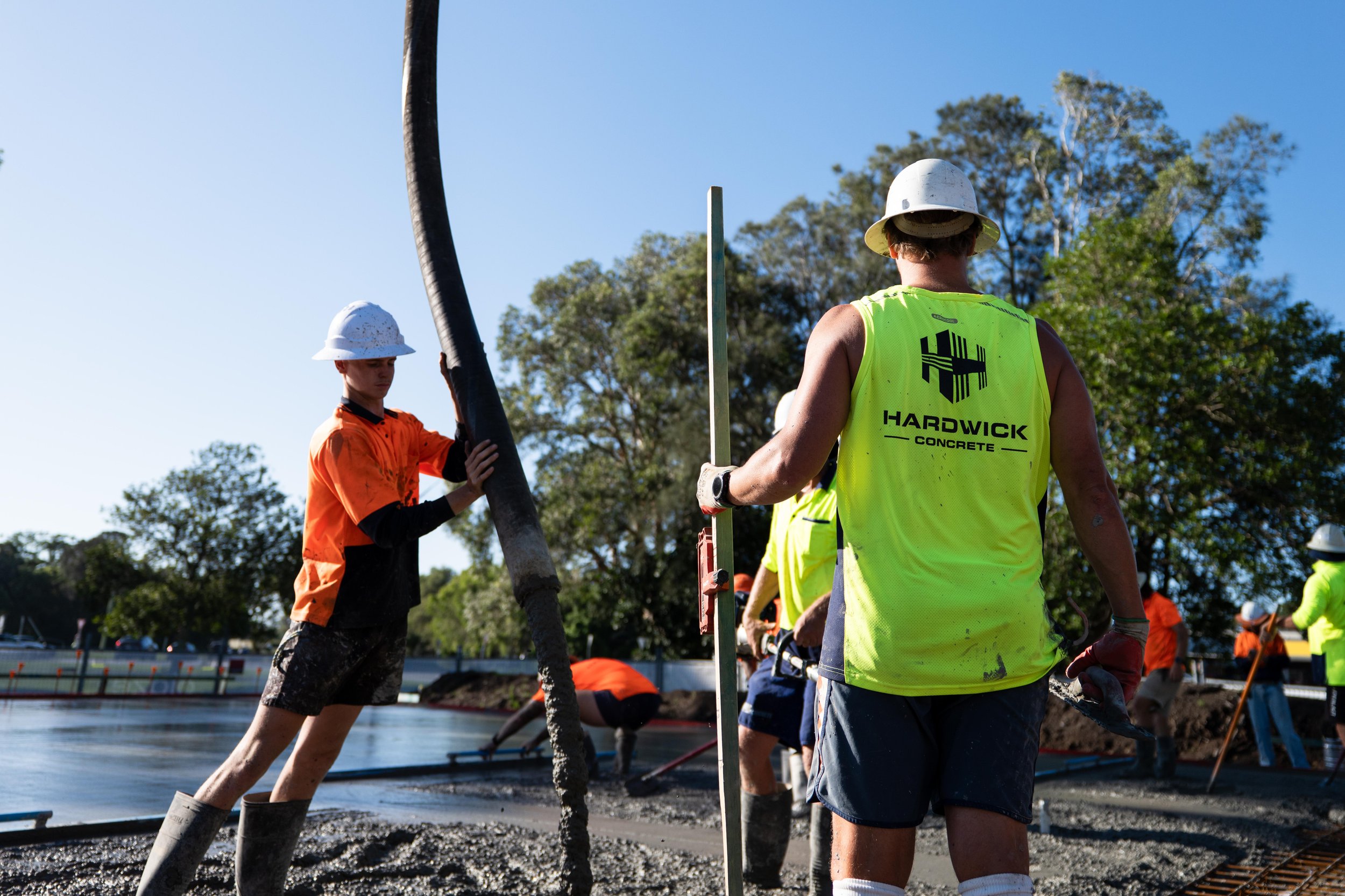 Construction workers wearing safety helmets and vests, pouring and leveling concrete on a foundation outdoors on a sunny day.
