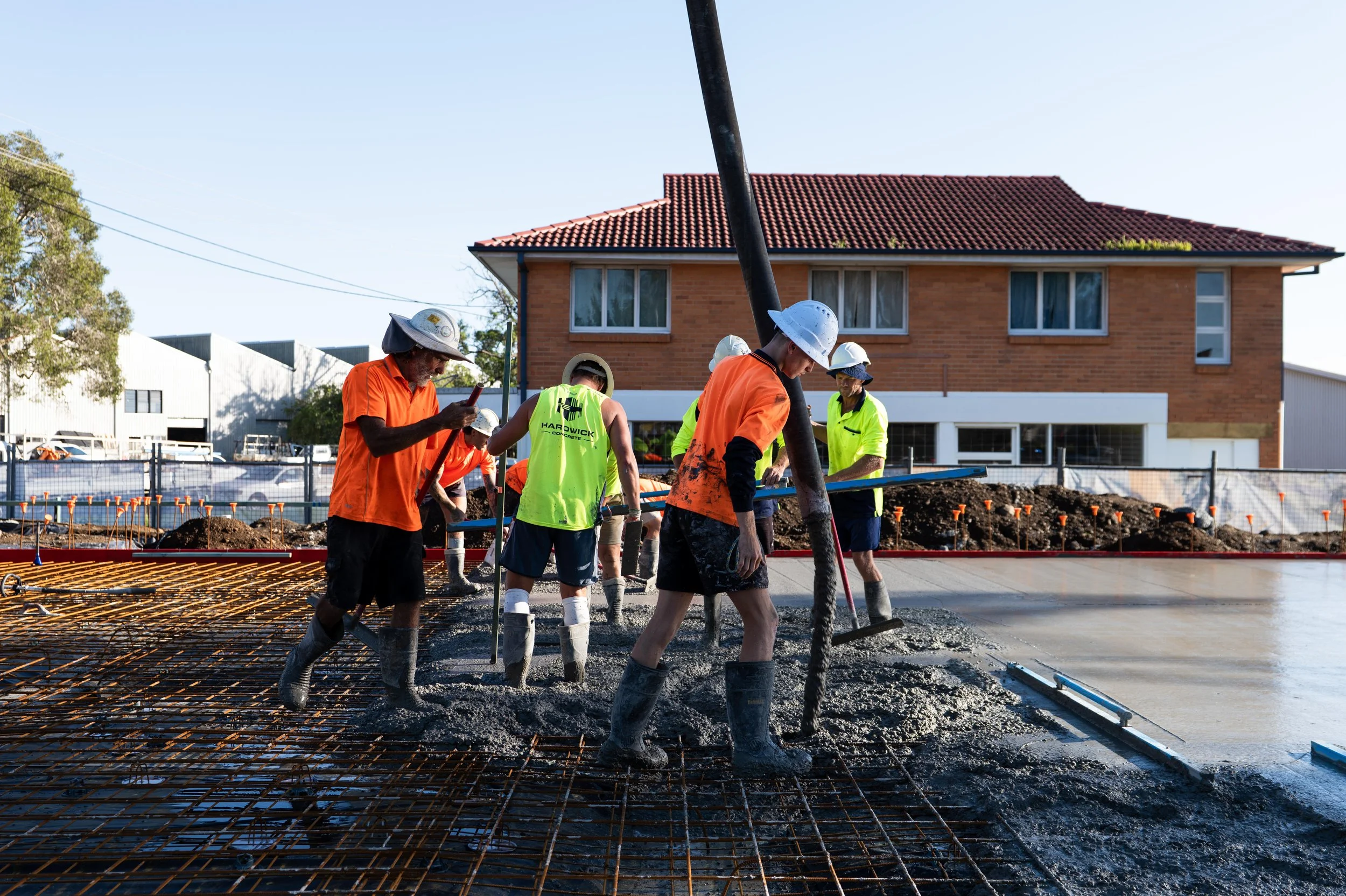 Construction workers pouring concrete on a building site with a house in the background.