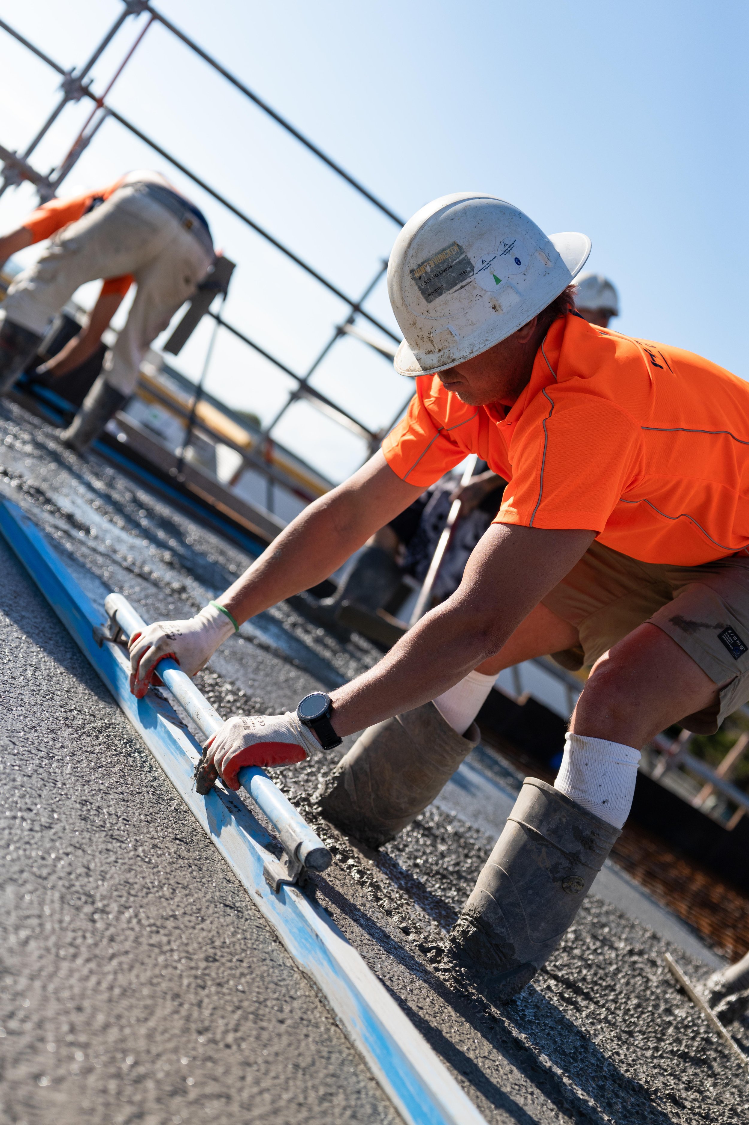 Construction worker in an orange shirt and white hard hat smoothing cement on a road.