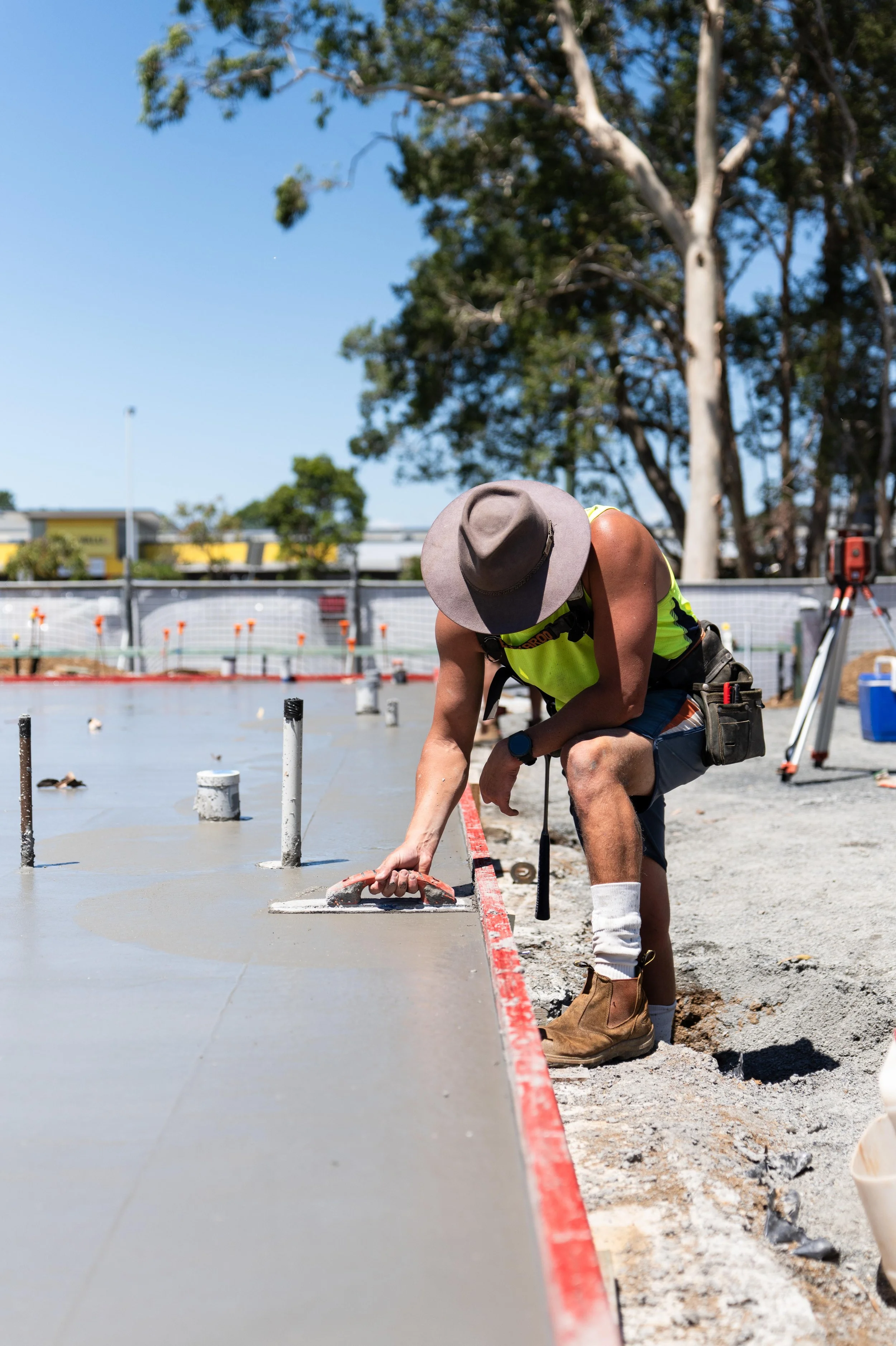 A construction worker wearing a gray hat, yellow safety vest, blue shorts, white socks, and brown work boots is working on a concrete slab outdoors during daytime. He is kneeling and smoothing the wet concrete with a hand trowel, with a background of