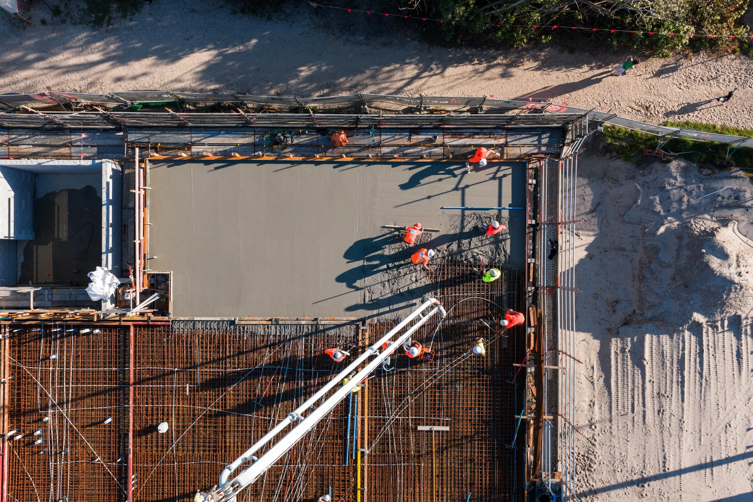 An aerial view of a construction site showing workers in safety vests and helmets installing a concrete slab, with gravel and dirt surrounding the area and construction materials and large pipes in the scene.