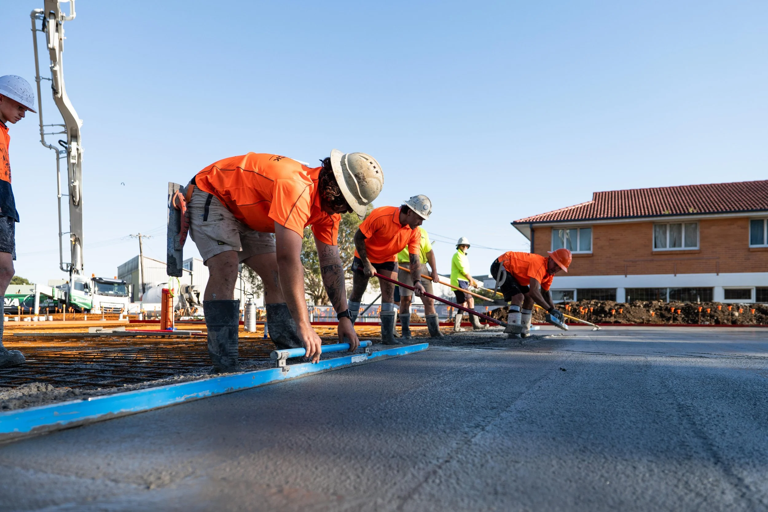 Construction workers in orange safety shirts and helmets smoothing wet concrete on a road under a clear blue sky.