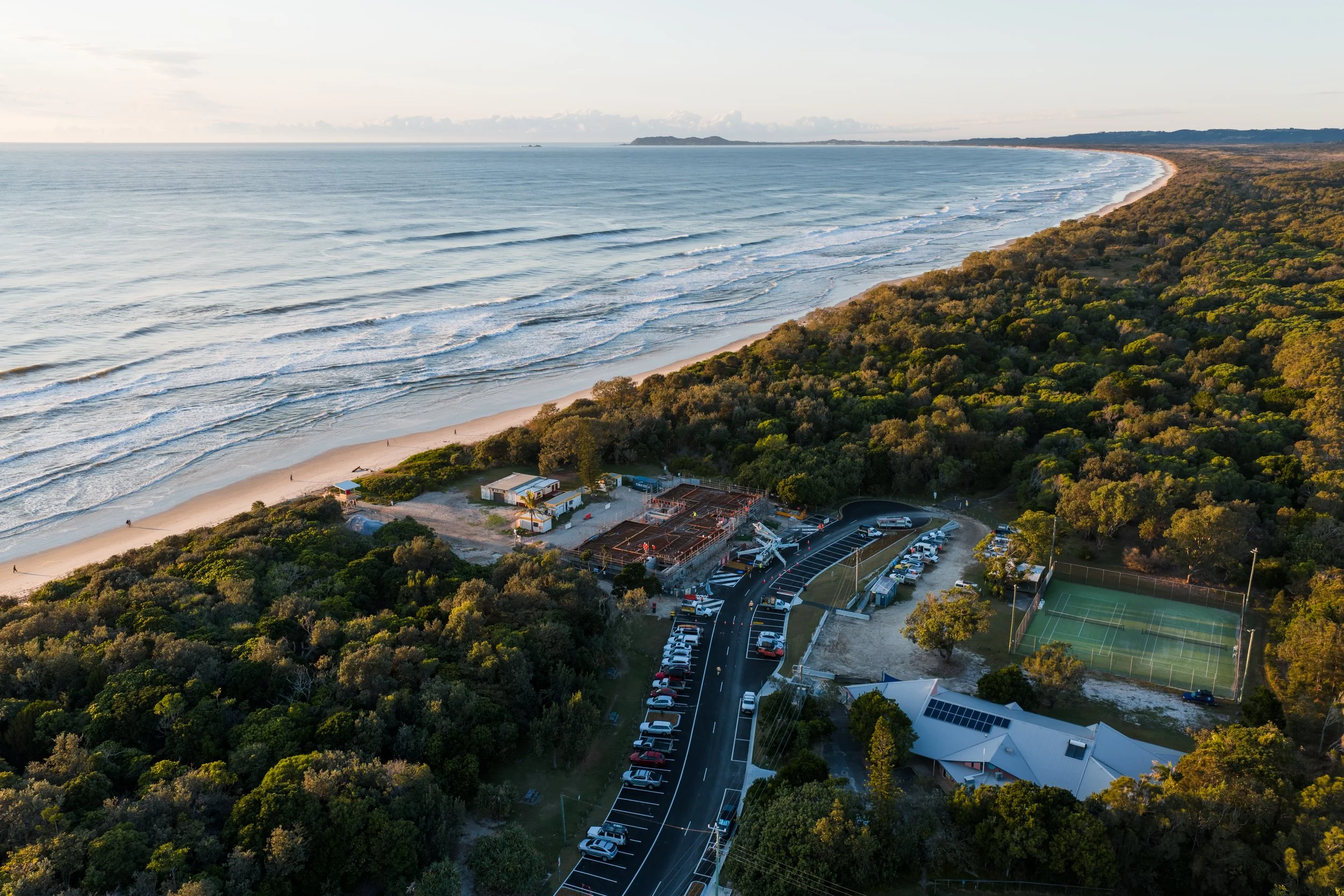 Aerial view of a beach with waves, a parking lot, tennis courts, a building, trees, and a construction site near the shoreline.