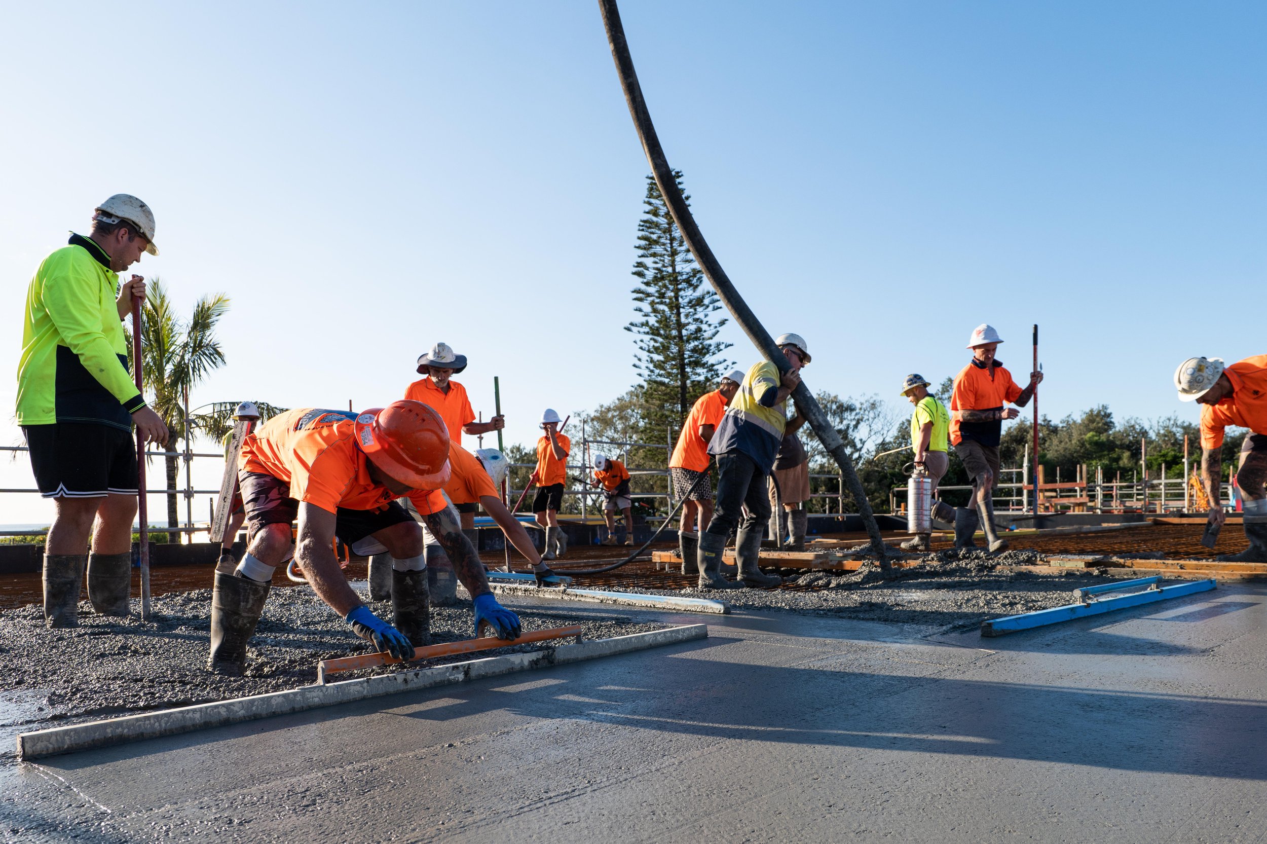 Construction workers in safety gear pour concrete on a construction site, with several workers leveling and smoothing the freshly laid concrete.