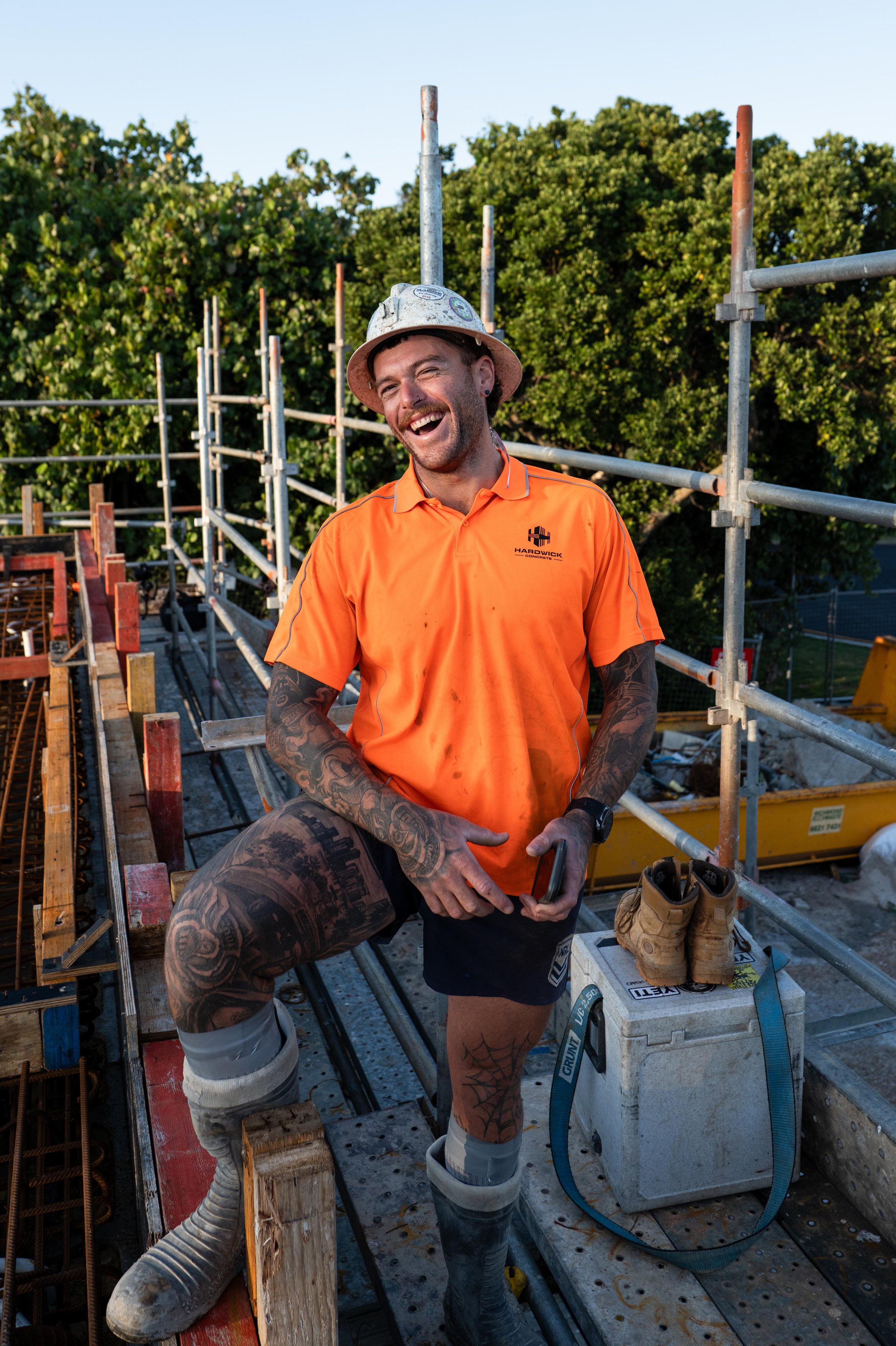 A smiling construction worker wearing an orange shirt, a hard hat, and tattoos, standing on a construction site with scaffolding and trees in the background.