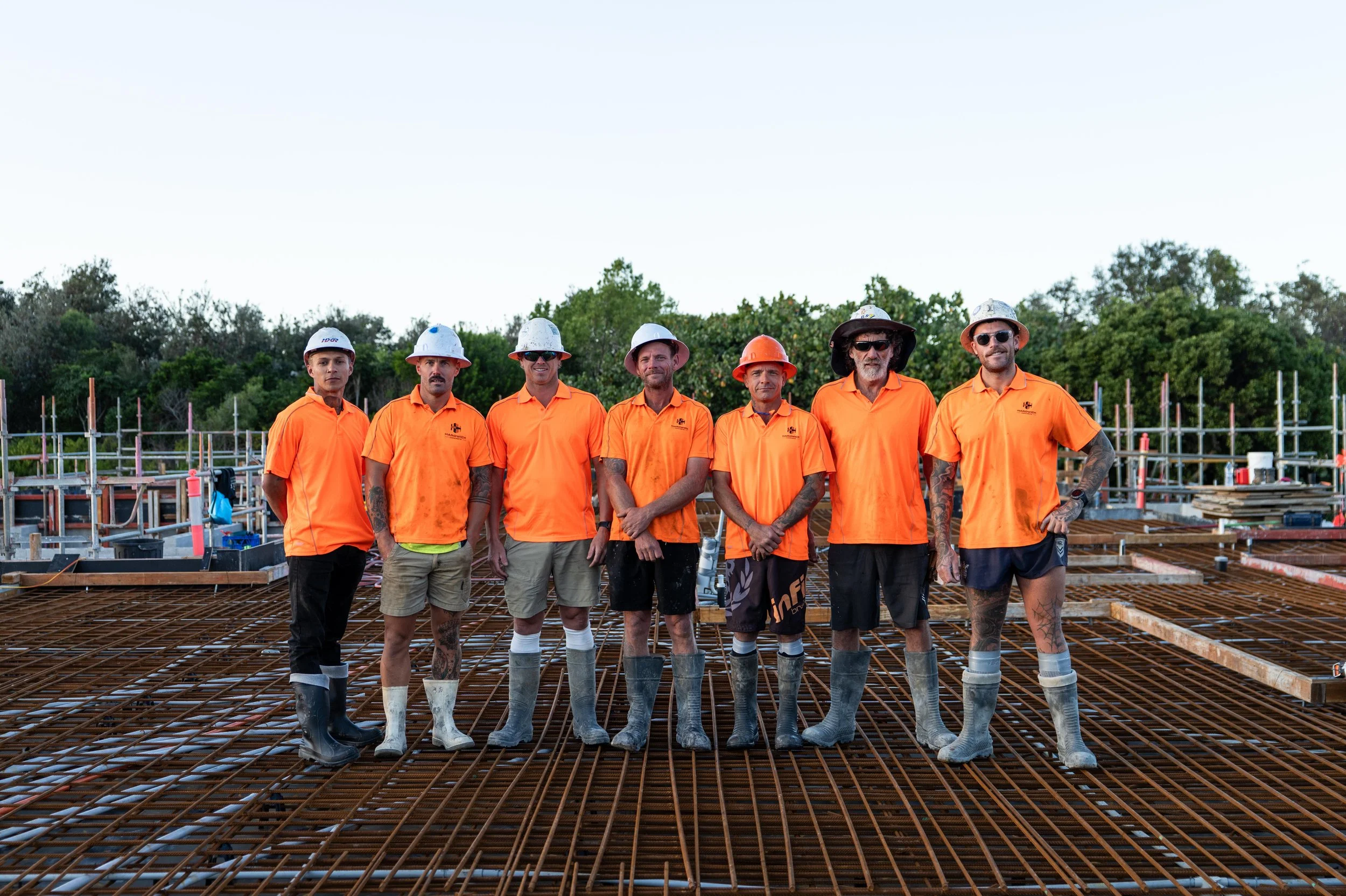 Construction workers standing on steel rebar at a building site, wearing orange shirts, hard hats, and rubber boots.