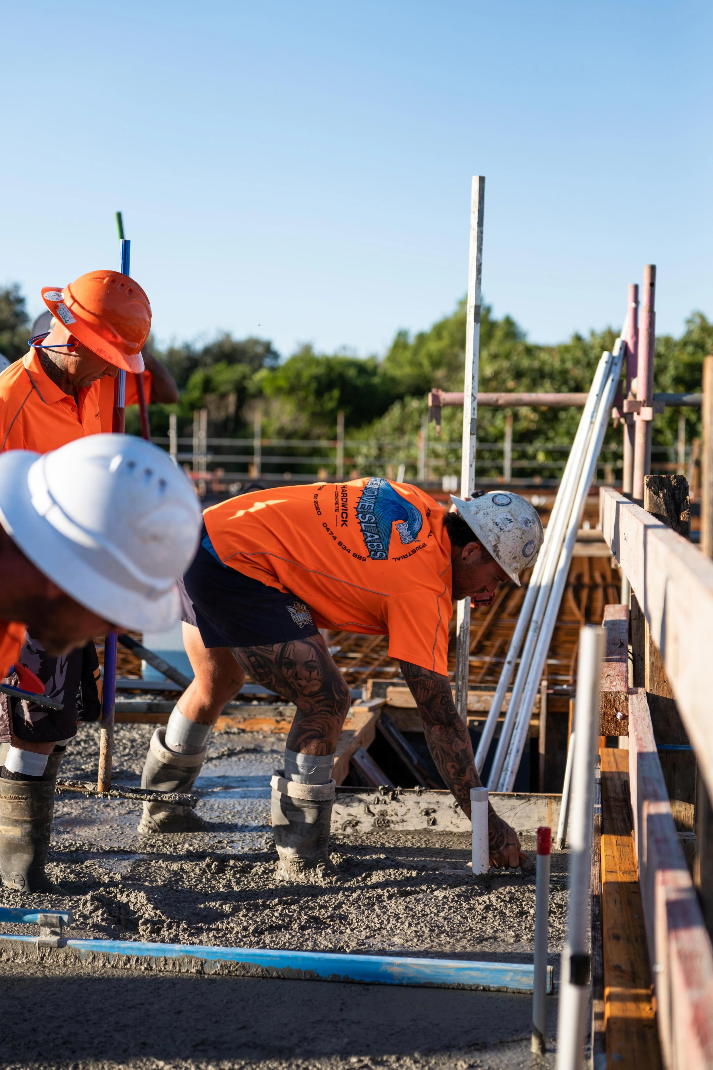 Construction workers in orange shirts and helmets pouring and leveling concrete on a building site during daytime, with trees and a clear blue sky in the background.
