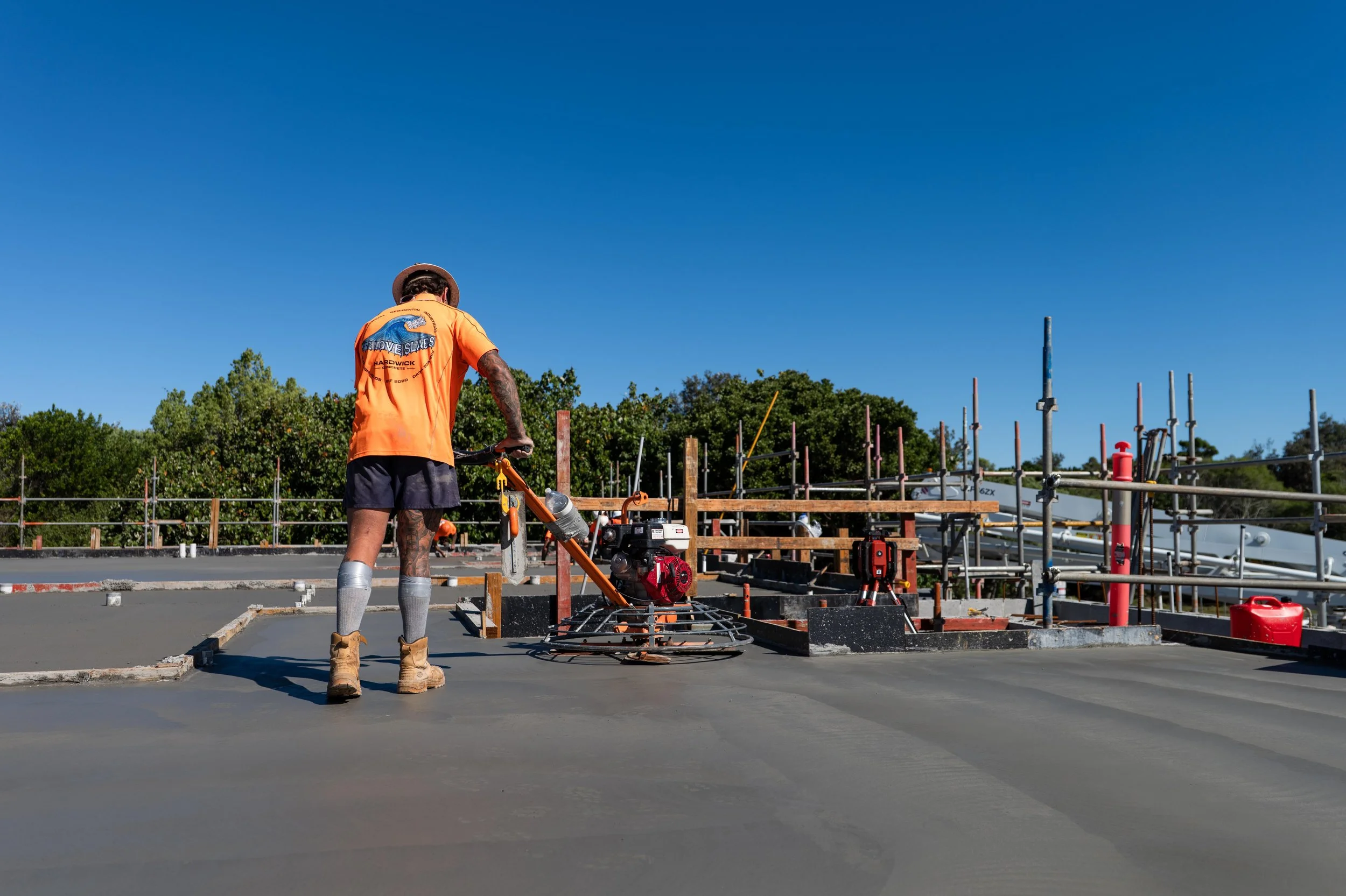 Construction worker in an orange shirt and boots smoothing fresh concrete on a building site under a clear blue sky.