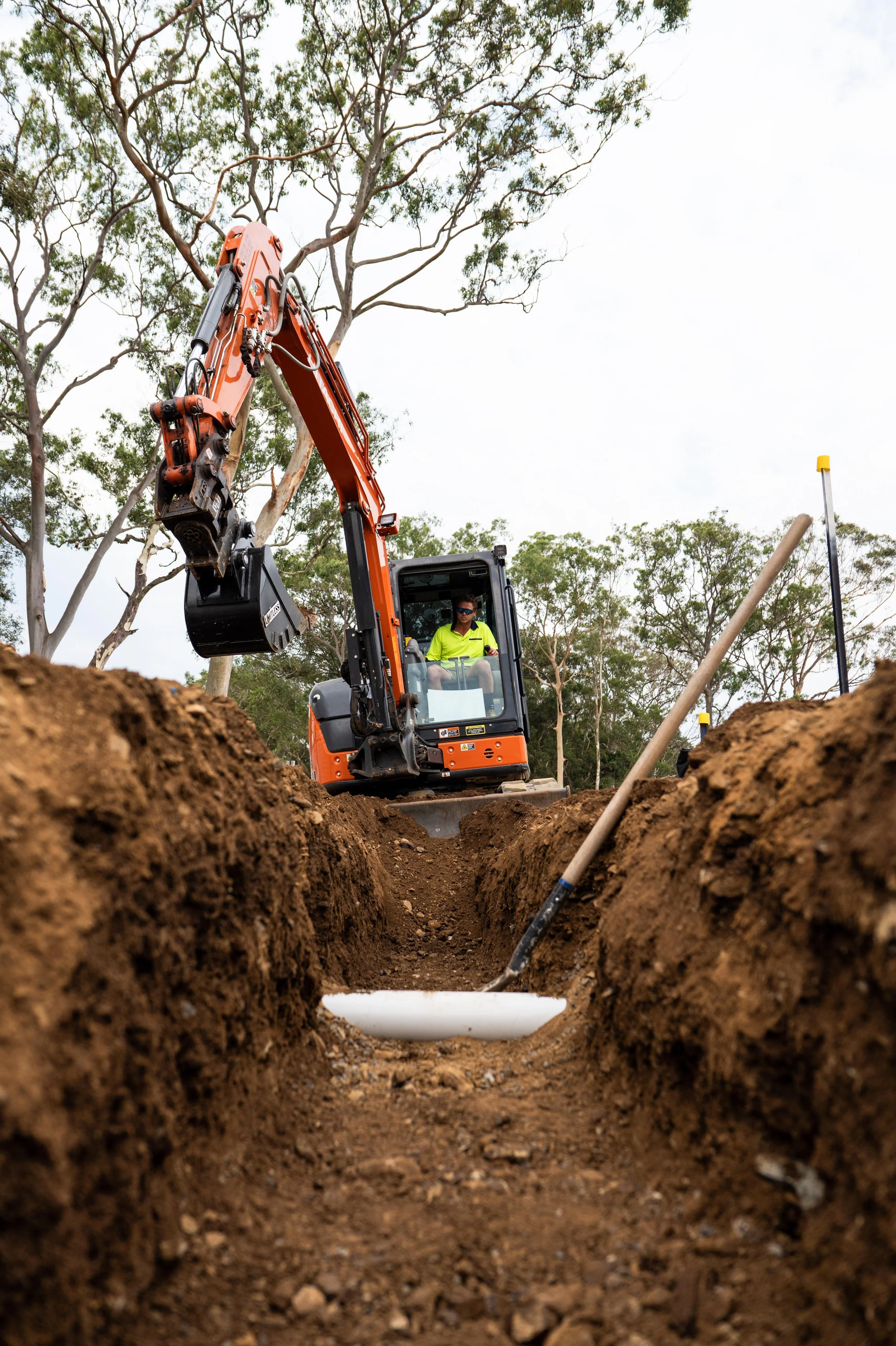 An excavator digging a trench in the ground at a construction site, with a worker in a bright yellow shirt operating the machine.