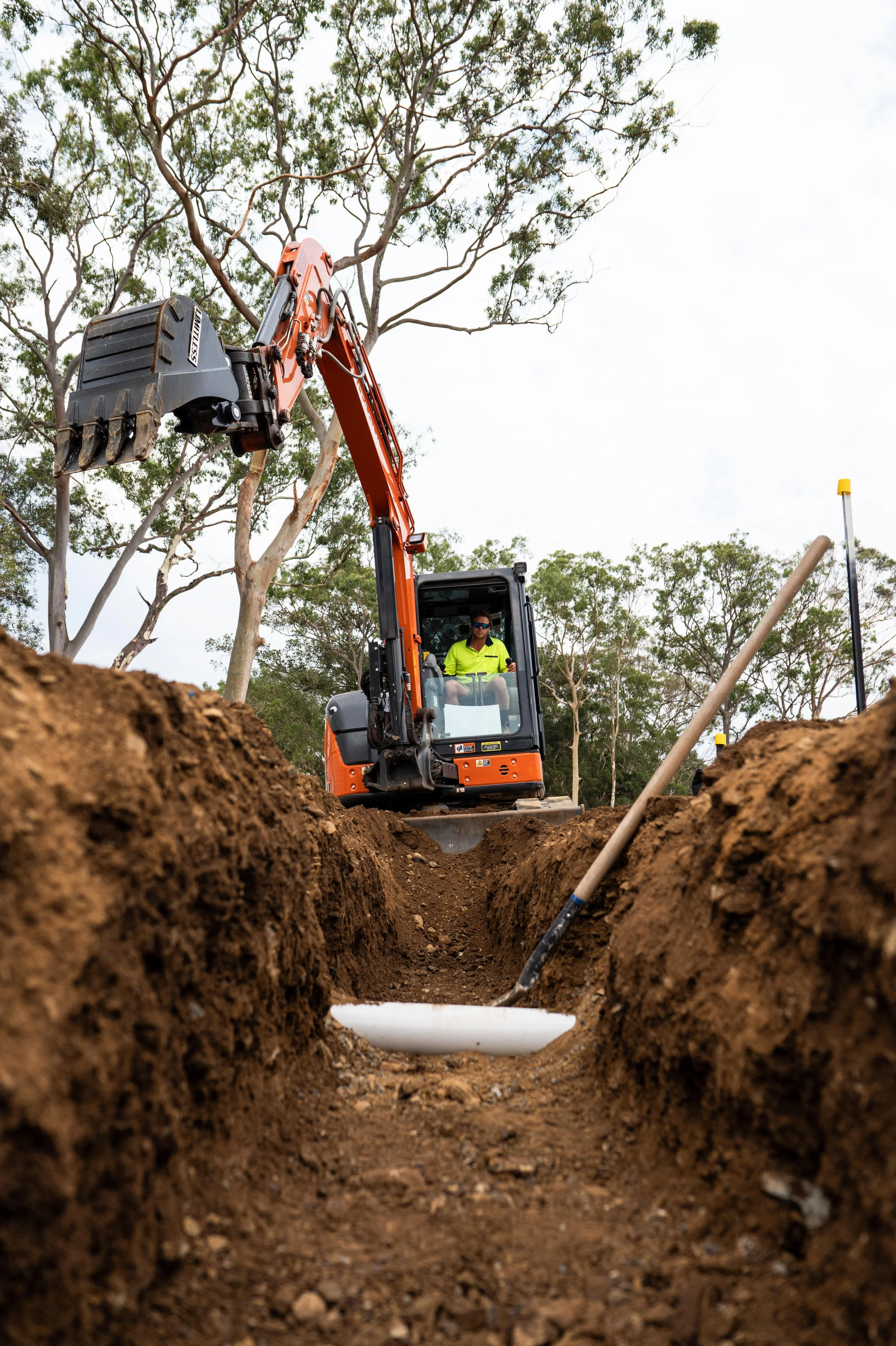 An excavator with a worker inside operating it, digging a trench in the ground outdoors with trees and cloudy sky in the background.
