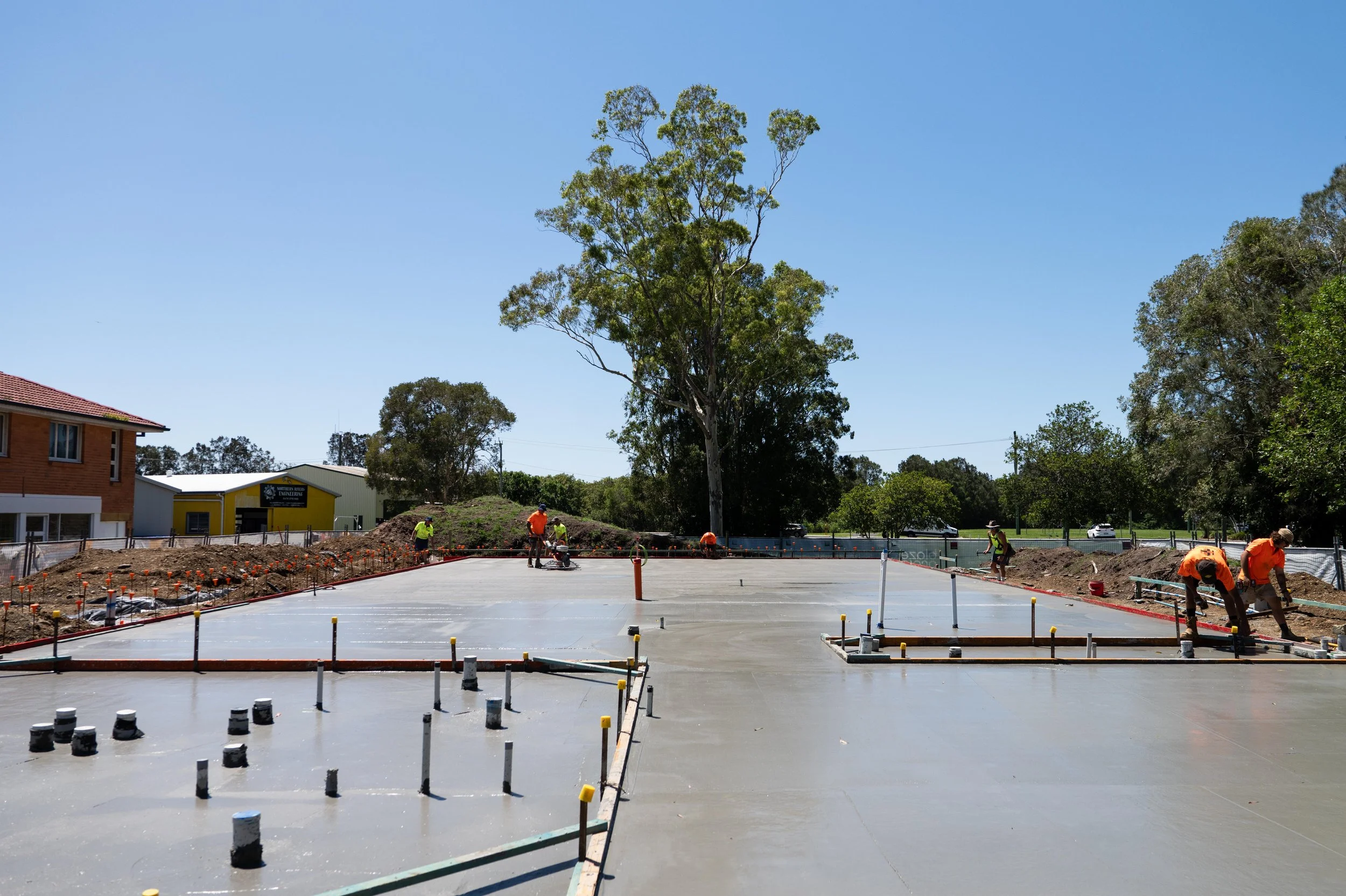 Construction workers leveling and smoothing freshly poured concrete on a building site with trees and buildings in the background.