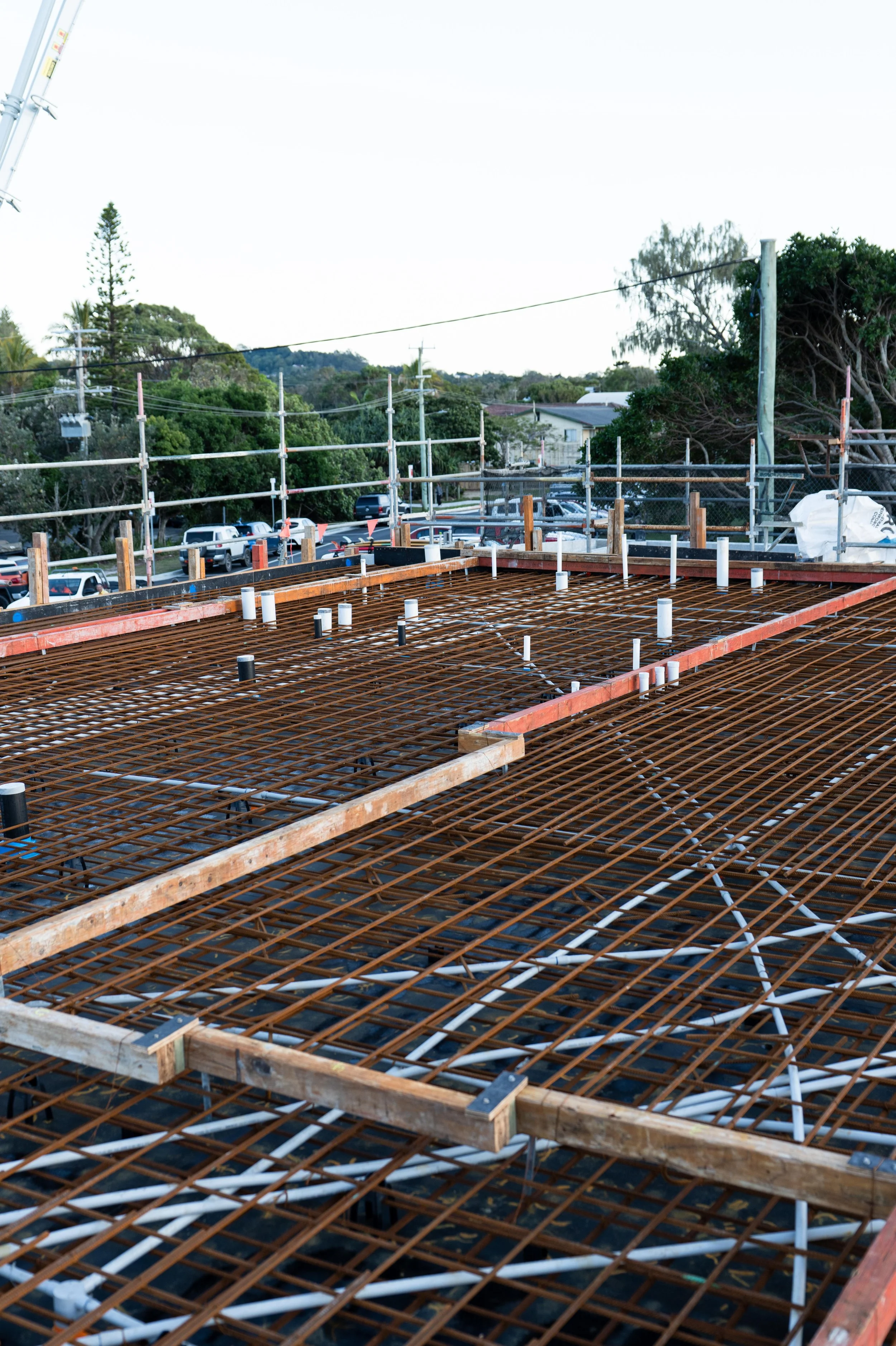 Construction site with rusted steel rebar for a building slab; white and black plumbing pipes protruding from concrete foundation; scaffolding and trees in background.