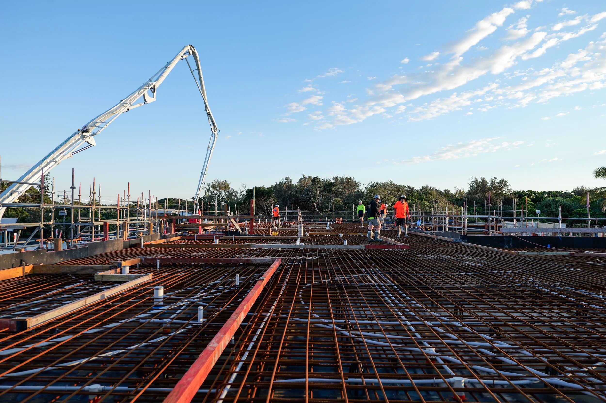 Construction workers on a building site with steel rebar, scaffolding, and a concrete pump in the background under a partly cloudy sky.