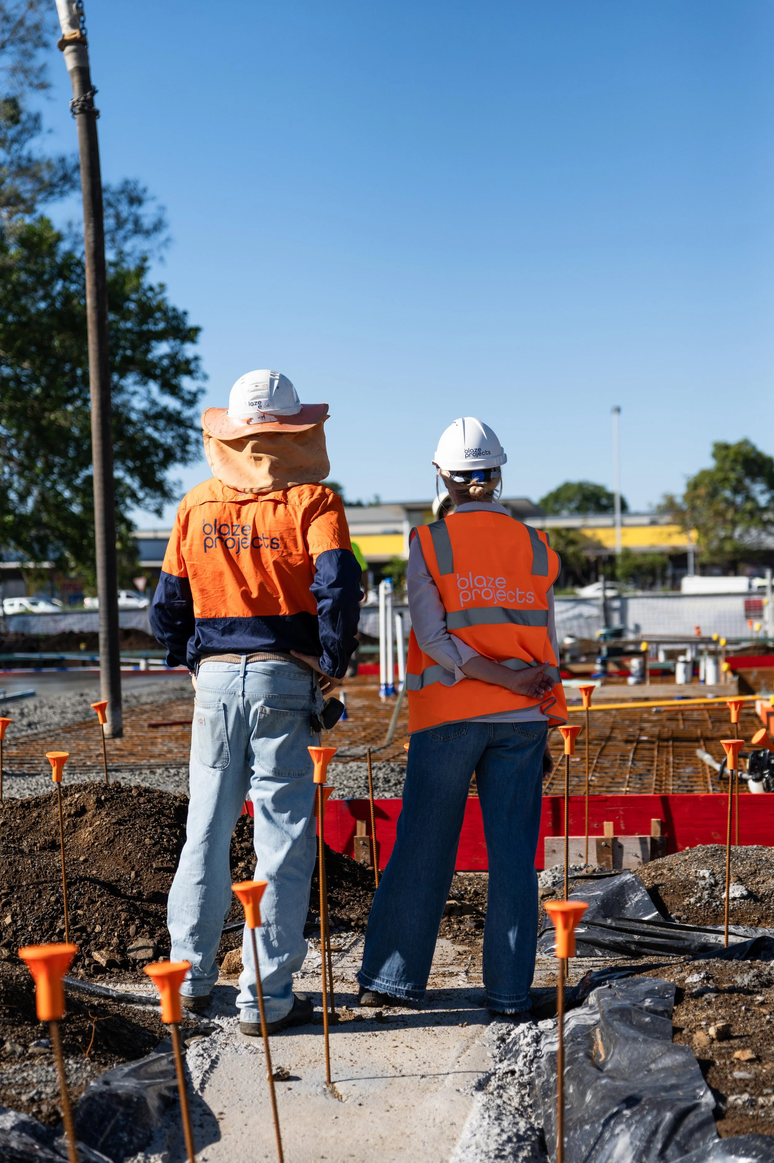 Two construction workers, one in an orange vest and the other in an orange jacket, stand on a construction site with their backs to the camera. They are wearing white safety helmets and are overlooking the site, which has dirt, construction markers, 