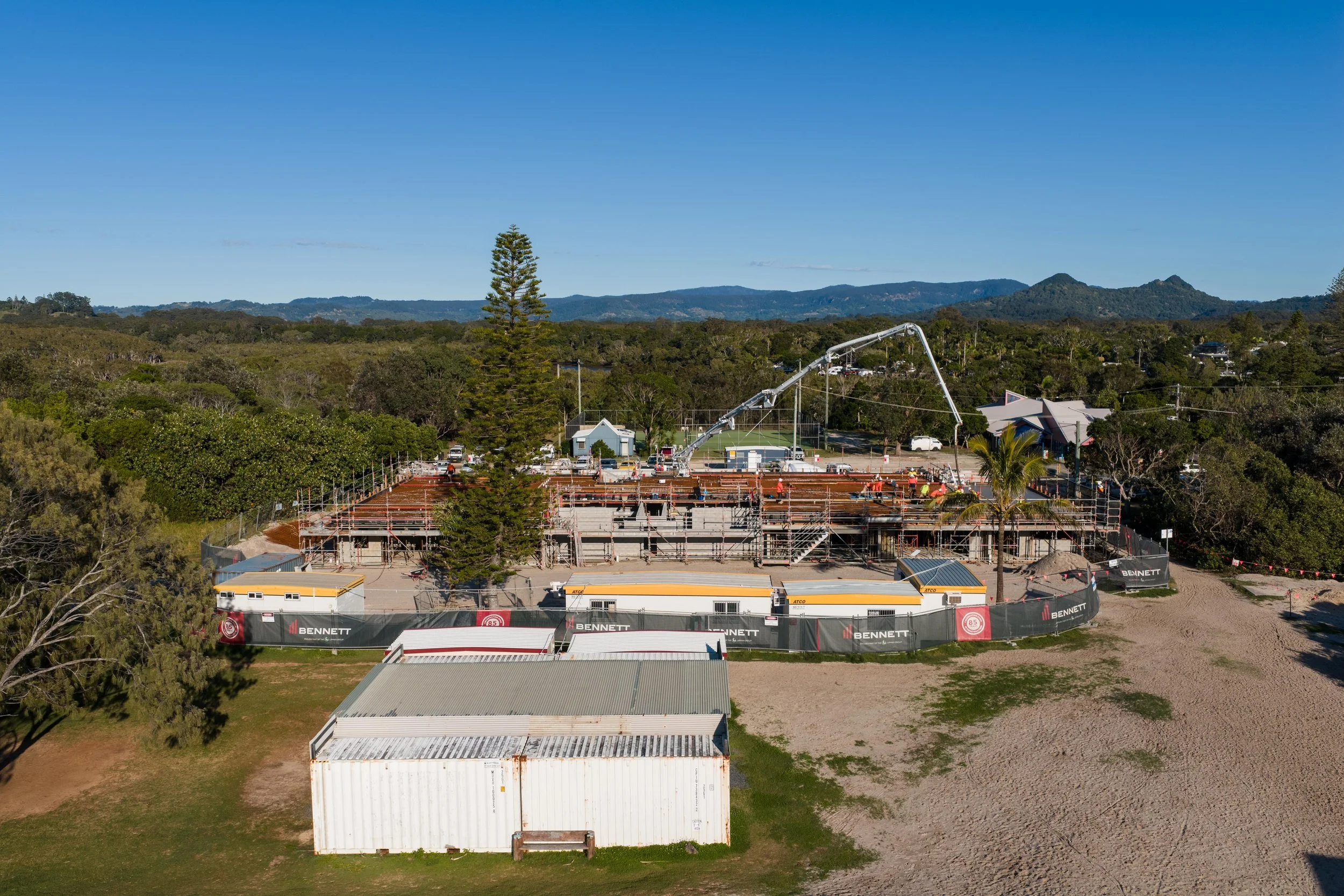 Under-construction building site with scaffolding, construction workers, and a concrete pump truck on a sunny day with trees and mountains in the background.