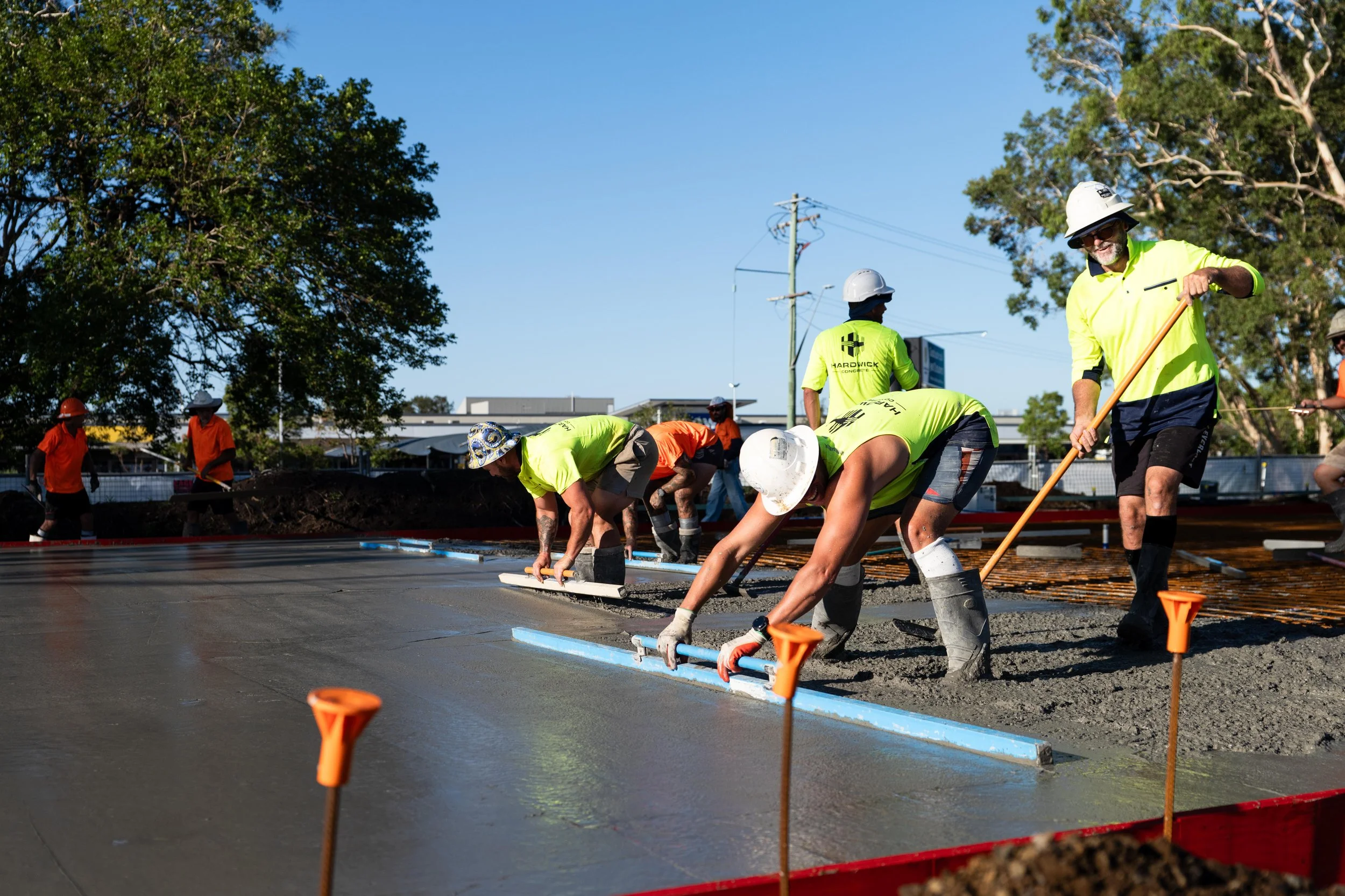 Construction workers pouring and leveling concrete on a sidewalk or road at an outdoor construction site with trees and utility poles in the background.